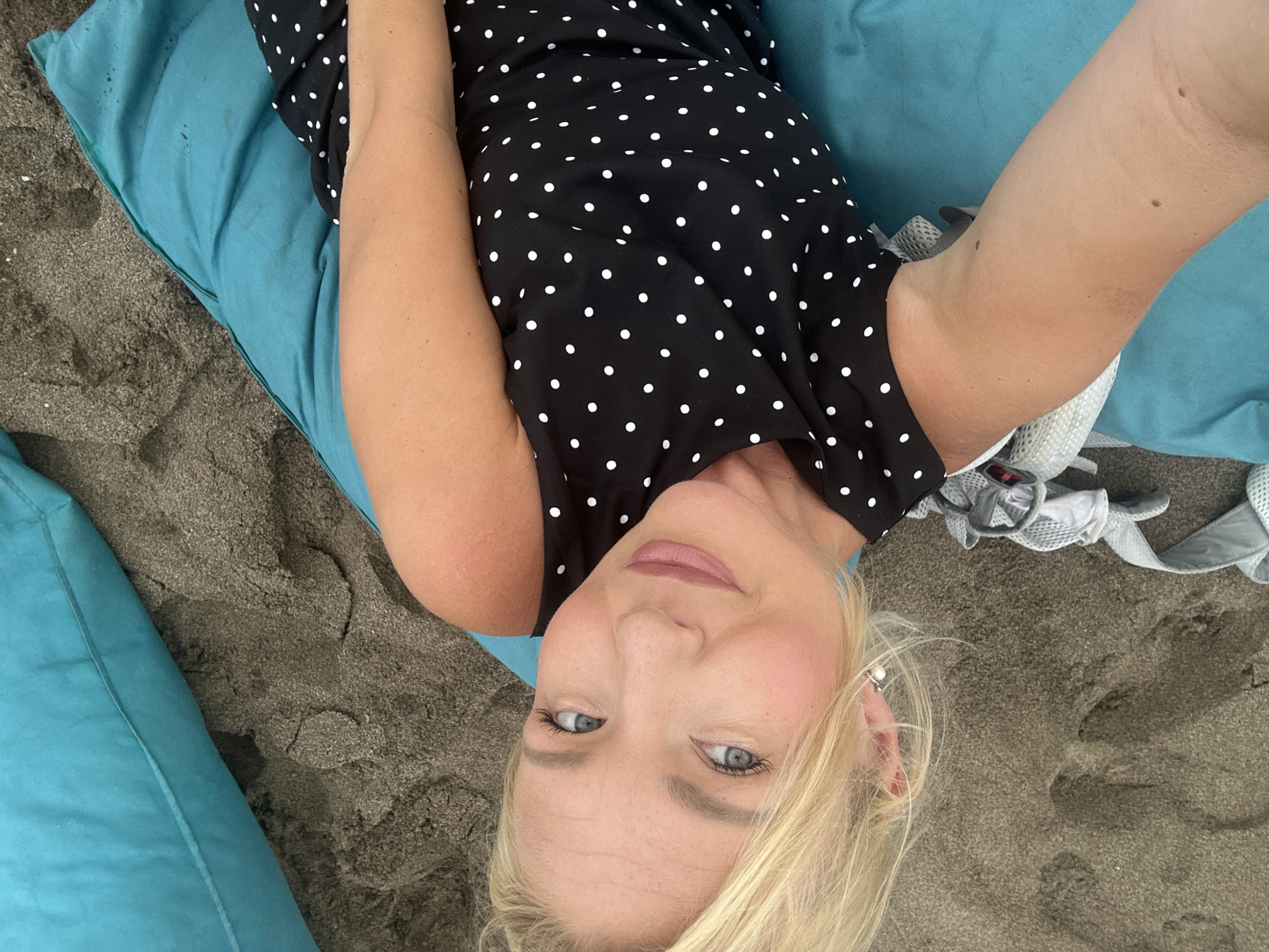 A blonde woman with blue eyes and fair skin is lying on a blue cushion on a sandy beach. She is wearing a black dress with white polka dots. Her left arm is extended towards the camera, and her right arm is bent, resting on the cushion. She is looking at the camera and smiling. The sand is dark gray, and there are some footprints visible. To the right of the woman, there is a large blue cushion. A light gray strap is visible in the sand to the left of the woman. The scene appears to be in North Kuta, Indonesia.