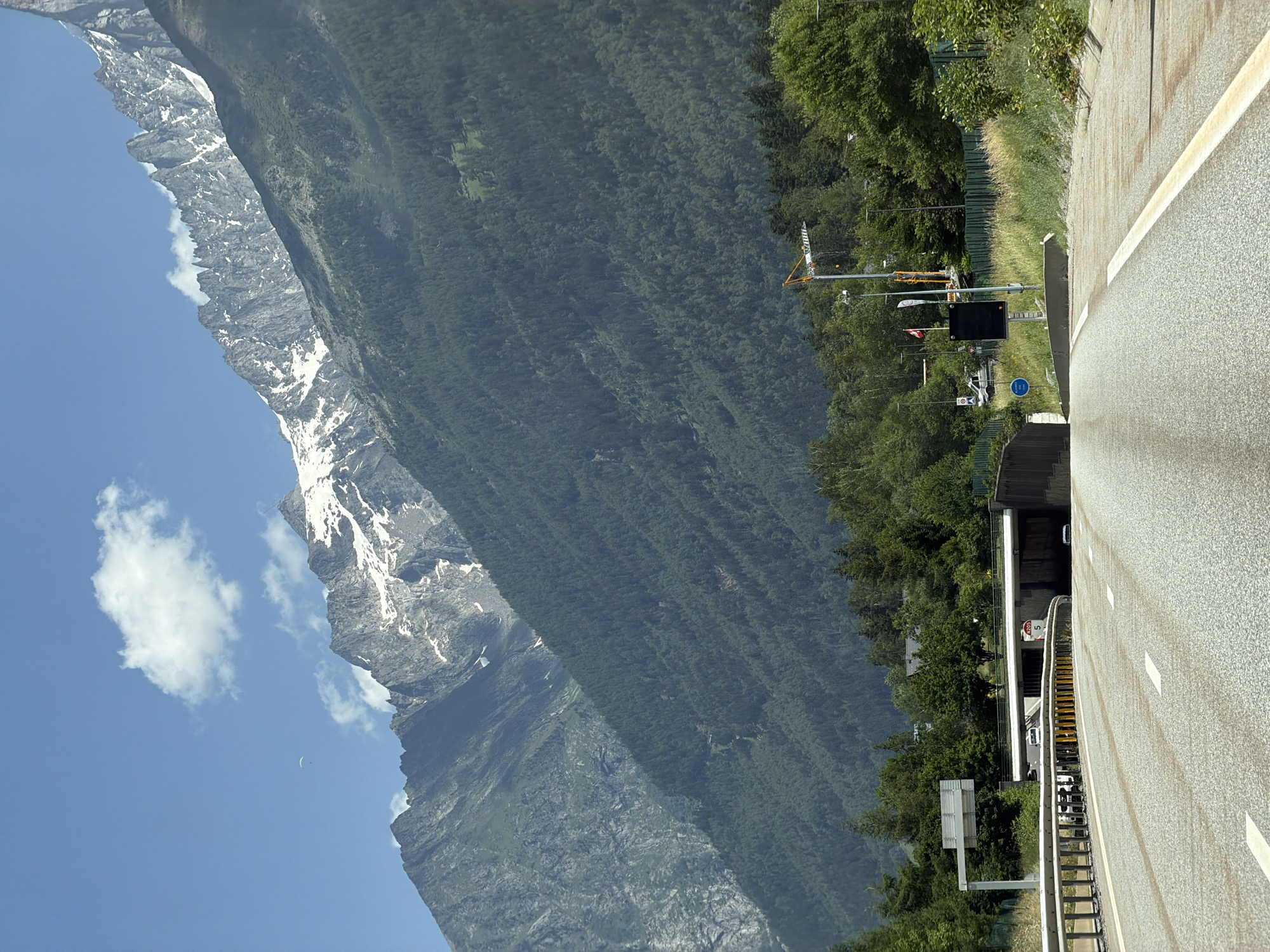 The image shows a highway leading towards a tunnel entrance in Chamonix-Mont-Blanc, France. Towering, snow-capped mountains dominate the background. The highway has white lane markings and guardrails. A paraglider is visible in the sky. In the middle ground, there are trees, buildings, and a construction crane. The sky is blue with a few white clouds.