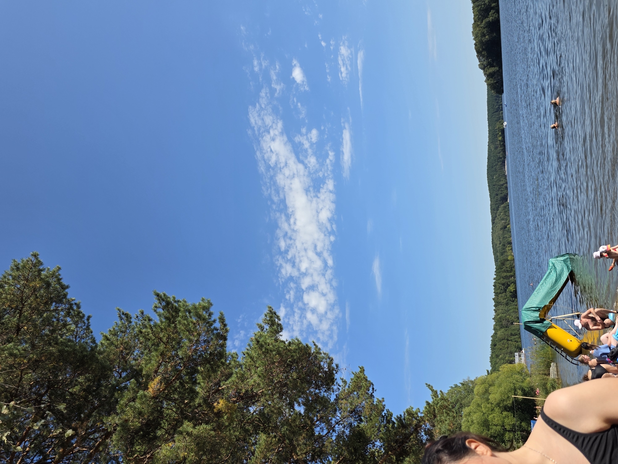 The image depicts a sunny day at a lake, likely in the city of Chebarkul, Russia, given the provided context. In the foreground, a woman's shoulder and upper back, wearing a black swimsuit, are visible on the left side of the frame. The beach area is populated with several people. To the right of the woman, a yellow water slide covered with a green tarp is partially submerged in the water, with a few individuals gathered around it. Some people are wading in the shallow water, while others are sitting on the sand. In the middle ground, the calm blue lake stretches out, with a few distant boats. Two people are swimming in the lake. The background features a dense, tree-covered shoreline, characteristic of the natural landscapes surrounding Chebarkul. The sky is a vibrant, clear blue with scattered white clouds. The overall scene captures a leisure activity on a warm summer day.