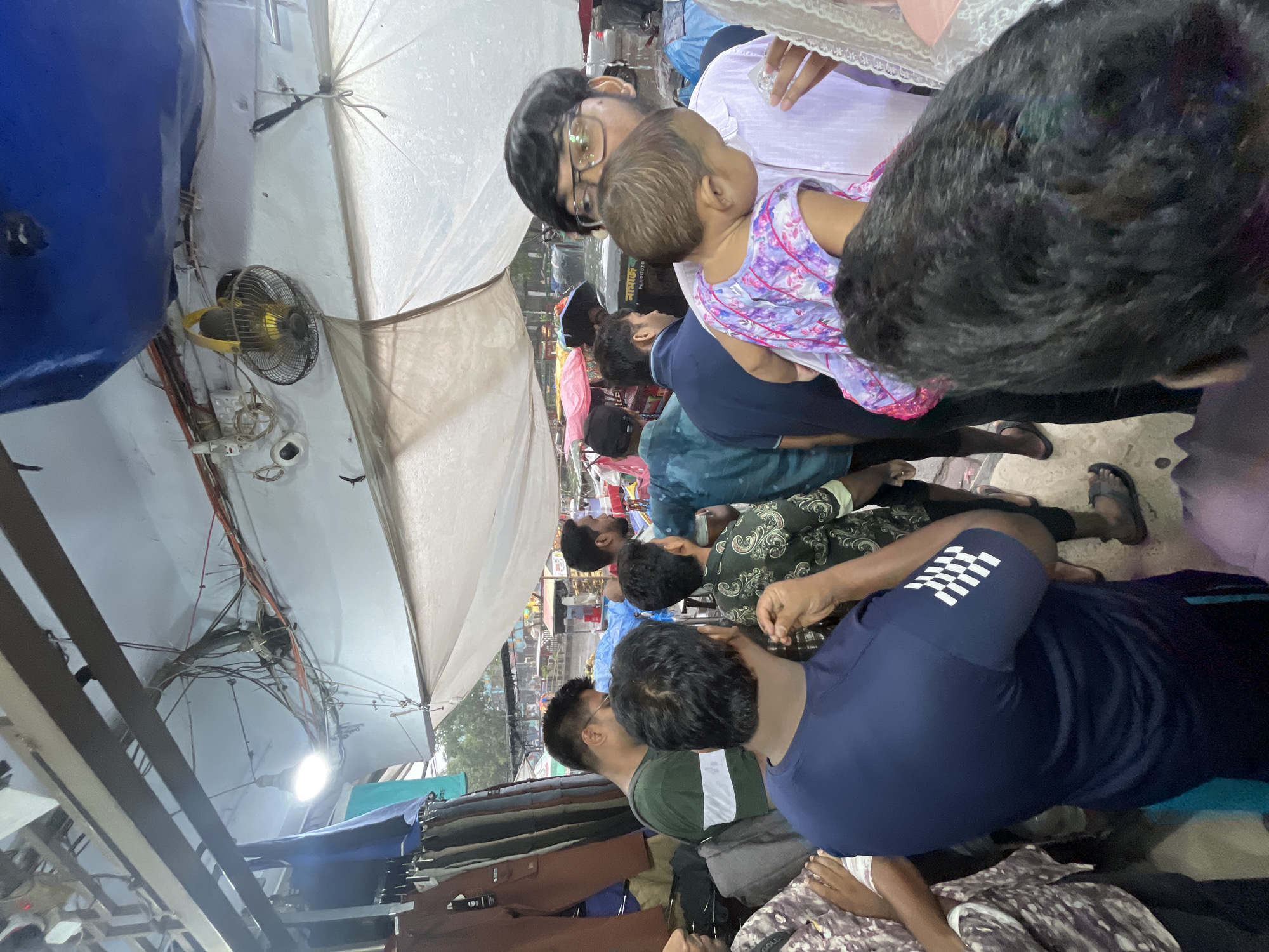 The image depicts an outdoor scene likely in a market area in Dhaka, Bangladesh. A group of men are gathered, mostly standing. One man is holding a child. They are under a canopy or awning, and electric wires are visible overhead. A small electric fan is attached to a support structure. Clothing items appear to be on display, suggesting a market or shopping area. The overall environment suggests a busy, potentially crowded urban setting.