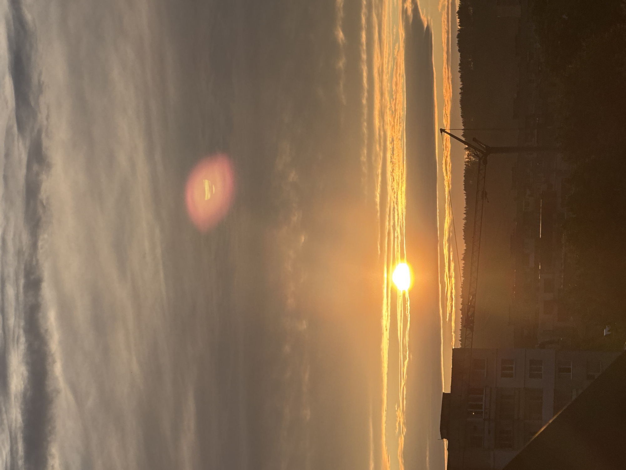 The image depicts a sunrise over the city of Pernik, Bulgaria. The sun, a bright orb of golden light, is partially obscured by horizontal clouds. A lens flare, appearing as a reddish-pink oval with an internal distorted image of the sun, is visible in the upper part of the sky. In the foreground, the silhouette of a multi-story apartment building is prominent on the left. Below and to the right, the landscape consists of more buildings and a dense tree line, with a construction crane silhouetted against the bright sky, suggesting ongoing development or infrastructure projects within the city. The overall atmosphere is one of a calm, early morning urban scene.