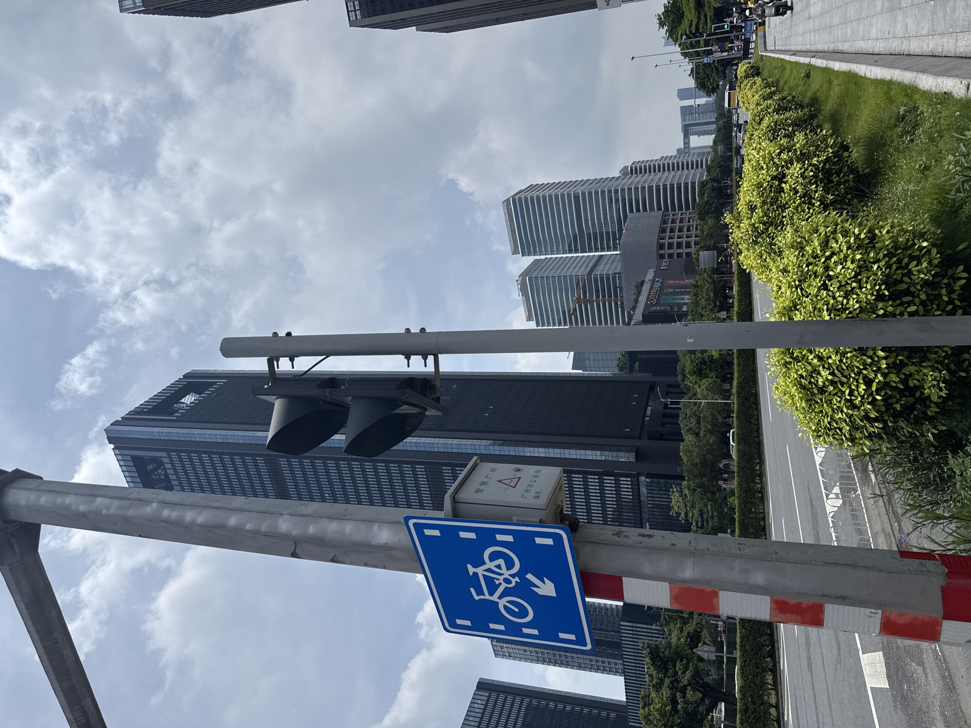 This detailed image depicts a bustling urban street scene in Guangzhou, Haizhu District, China, under an overcast sky with diffused white clouds.
In the immediate foreground on the left, a robust gray pole, marked with alternating red and white reflective stripes at its lower section, extends vertically out of the frame. Attached to this pole is a square blue sign with rounded corners, featuring a white pictogram of a bicycle positioned above a downward-pointing arrow, indicating a designated bicycle route. Directly beneath this sign is a small, light gray metal utility box. Chinese characters on the box read "智慧广州" (Smart Guangzhou) at the top, and "广州市公安局 编号:" (Guangzhou Municipal Public Security Bureau, serial number:) below, confirming the location within Guangzhou.
To the right, another gray metal pole holds a black traffic light, which is oriented away from the viewer. A well-maintained median strip, composed of neatly trimmed, vibrant green, small-leafed bushes, runs parallel to the road. Beyond this, a multi-lane asphalt road with clear white lane markings extends into the distance, accommodating moderate vehicle traffic, including several cars. White metal guardrails are visible along the edge of the median strip. On the far right, a gray paved sidewalk is present, where several individuals on motorcycles or scooters are either parked or slowly navigating.
The background is dominated by a dense array of modern high-rise buildings. A prominent tall, dark skyscraper with a grid pattern of windows and a distinctive open section near its top stands on the left. To its right, a cluster of other contemporary high-rise structures, some characterized by distinct horizontal architectural bands, contributes to the skyline. In the distant center, an iconic arch-like building, appearing to connect two towers, is discernible against the cloudy sky. Lush green trees are scattered throughout the scene, interspersed between buildings and along the edges of the road, adding natural elements to the urban environment.