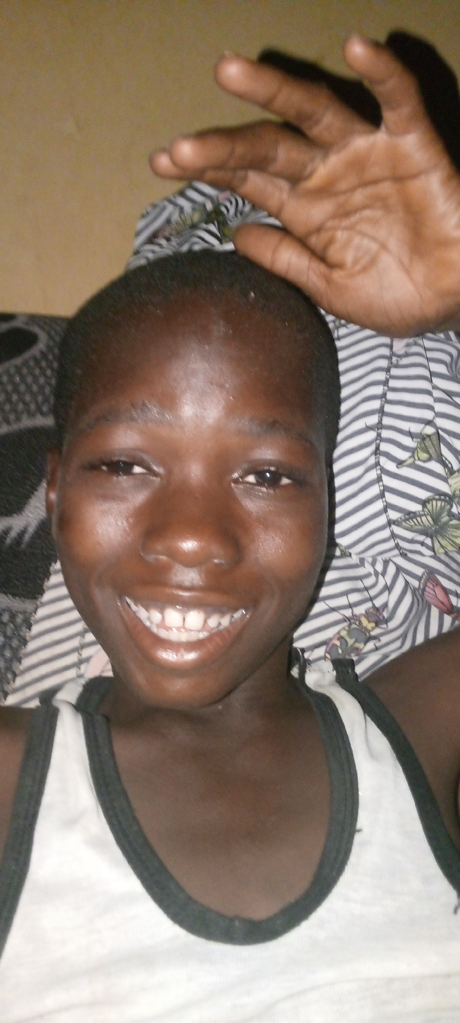 A close-up of a smiling young Black person in a white tank top with black trim. The person's hand is positioned near the top of their head. The person is laying on a bed. The bedsheets are black and white, and the background wall is a plain beige. The location appears to be indoors, but specific details to confirm it being Adankolo, Nigeria, are not available in the image.