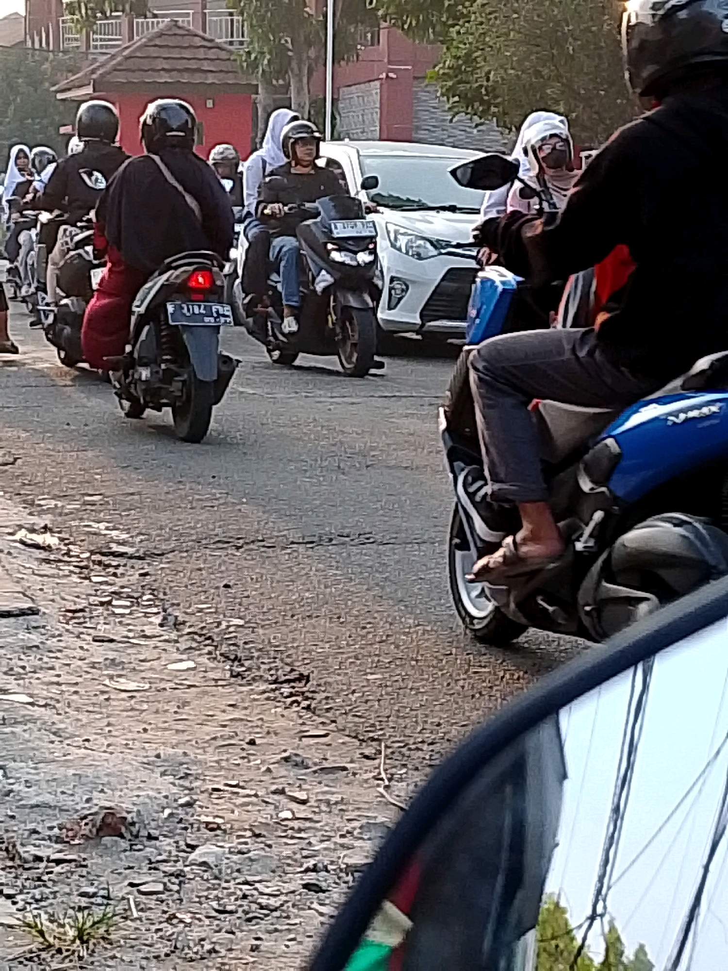 The image captures a street scene in Cibinong, Indonesia, likely during daylight hours, with a focus on multiple individuals riding motorcycles. In the foreground, a person on a blue motorcycle is visible from the waist down, wearing dark pants and a sneaker on their left foot, with their right foot resting on the motorcycle's footboard. The motorcycle has a dark-colored seat and blue body panels, and part of its front fairing is visible. Further back, a line of motorcycles and riders are navigating a paved road with some visible cracks and patches.
Several individuals are wearing helmets, including one on a black motorcycle in the middle ground whose rider is wearing a black jacket and red pants. Another rider on a similar motorcycle ahead of them is wearing a helmet and dark clothing, with a white object on the back of their seat. To the left, a white car is present, and several more people on motorcycles are visible further down the street. The background shows a building with a reddish-brown tiled roof, trees, and power lines, suggesting an urban or semi-urban environment. The overall atmosphere appears to be one of daily transit, with multiple vehicles sharing the road.