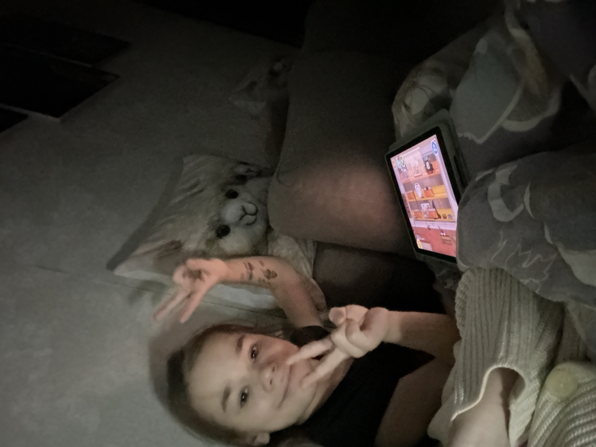 The image shows a young person, likely a child or pre-teen, lying down in a dimly lit indoor setting. They are looking directly at the camera with a subtle smile and are making peace signs with both hands, suggesting a relaxed and playful mood.
To the left of the person's head, partially obscured by a pillow, is a fluffy white stuffed animal resembling an alpaca or sheep. A tablet computer, encased in a light-colored protective cover, is positioned upright to the right, displaying a colorful, cartoon-style game on its screen. Small icons and numbers, including "00G" and "000" at the top left and a circular "!" icon at the top right, are visible on the tablet.
The setting appears to be a cozy indoor space, possibly a living room or bedroom, furnished with soft, grey-toned bedding or couch cushions. The dim lighting implies it is evening or night. A light-colored, ribbed knitted fabric, possibly a blanket or clothing, is visible on the right side of the frame, covering part of the person's lower body. This scene is set in Uva, Russia.