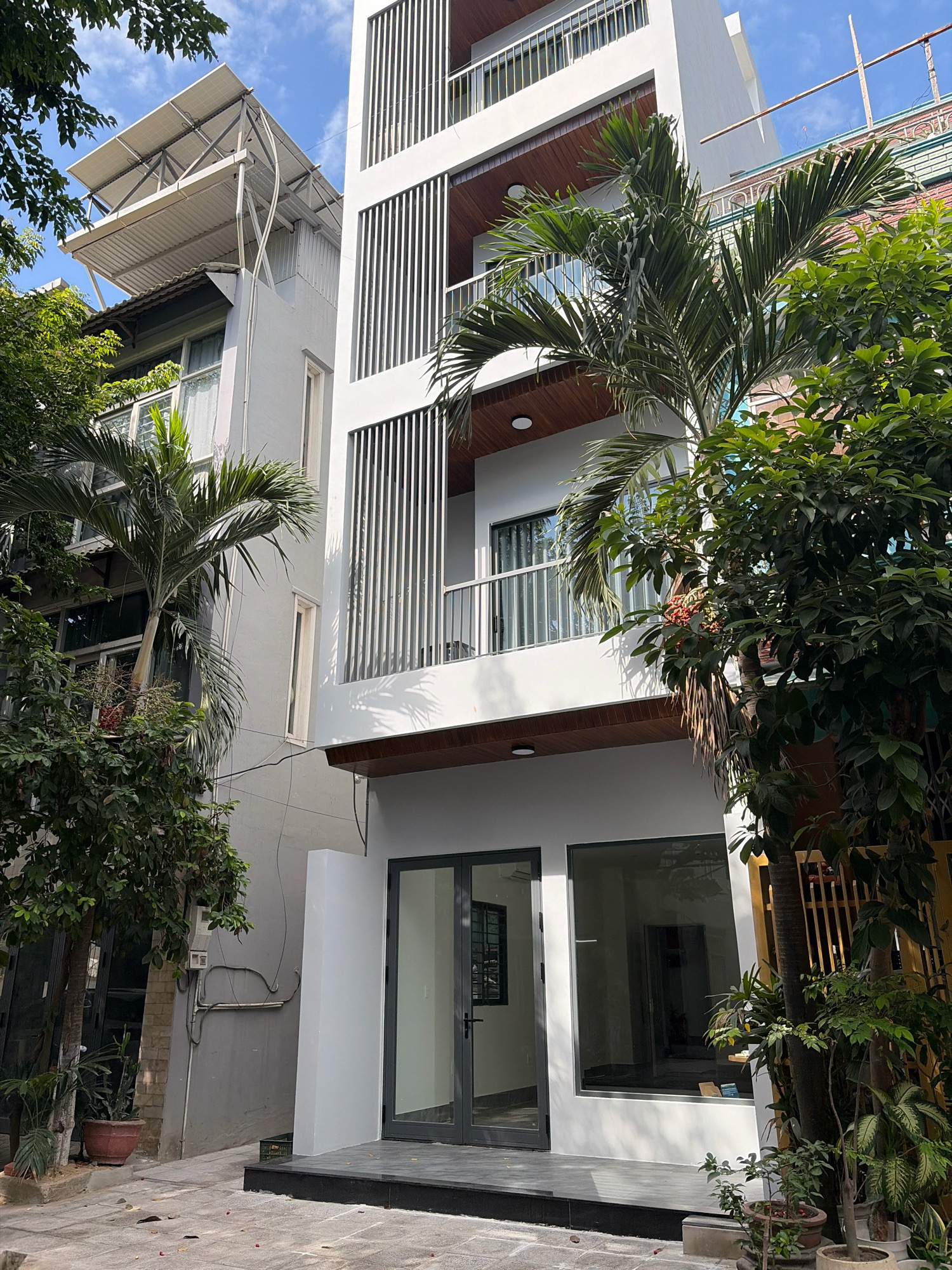 The image captures a modern multi-story building in Đà Nẵng, Vietnam, with a bright blue sky and white clouds overhead. The building features a white facade with vertical slats on the upper floors, balconies with white railings, and dark wood accents. Solar panels are installed on the roof of an adjacent, older building to the left, which has a more traditional aesthetic with a grey facade and windows with white curtains. Lush green trees and palm trees with large fronds surround the buildings, with some trees bearing small, reddish-brown fruits. In the foreground, the ground is paved with grey and black tiles, and there are several potted plants. The ground floor of the modern building has large glass doors and windows. The scene appears to be a quiet street during the daytime.
