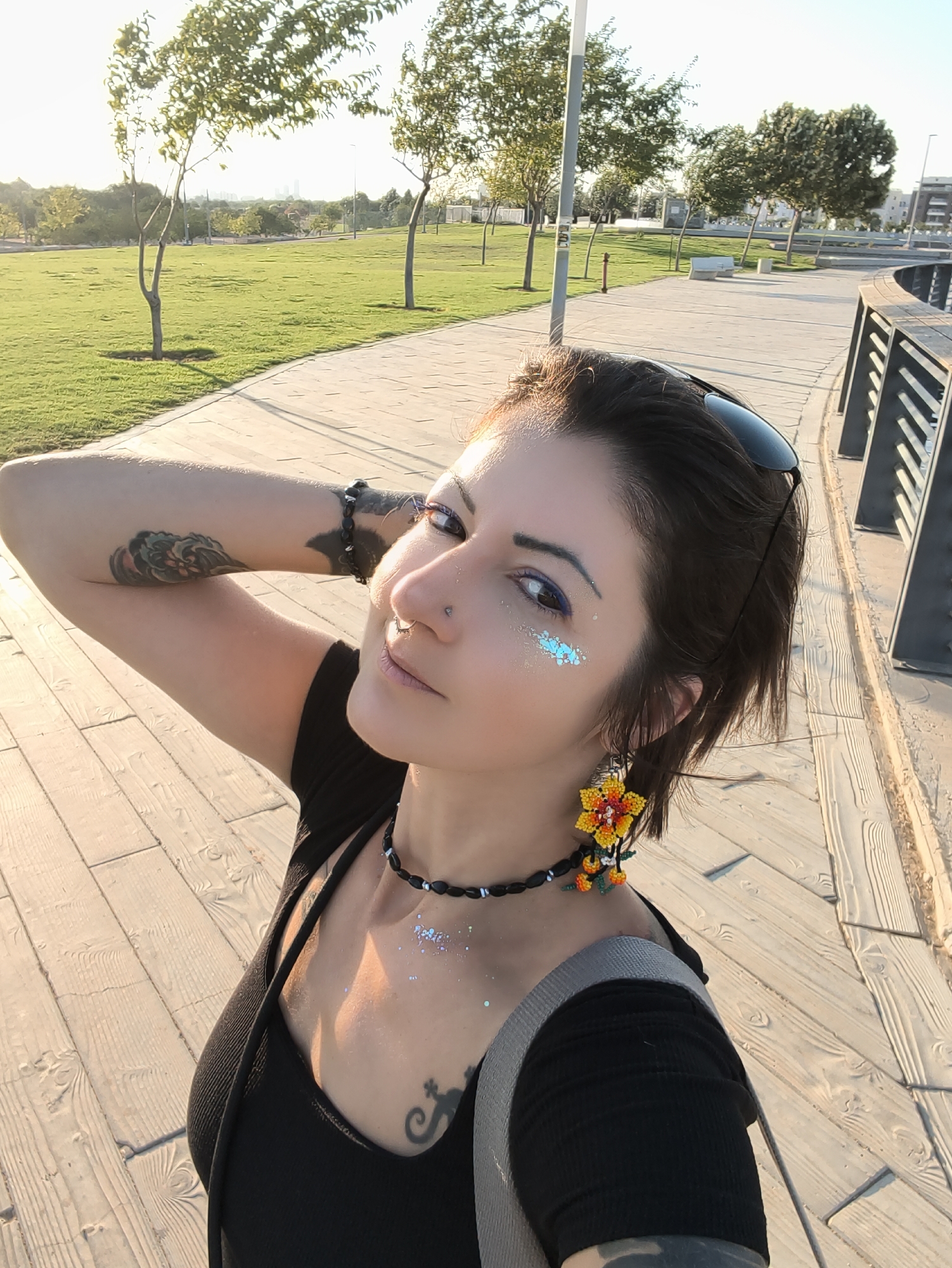 A young woman with dark hair, sunglasses perched on her head, and artistic makeup including blue eyeliner and glitter on her cheekbones, poses for a selfie outdoors. She is wearing a black top, a beaded necklace, and a colorful beaded earring. She has tattoos on her arm and chest. The background features a wooden boardwalk, a grassy park with trees, and a city skyline in the distance. This scene is likely in the area of Be'er Sheva, Israel, possibly near the Beer Sheva River Park, a common urban green space with boardwalks and recreational areas.