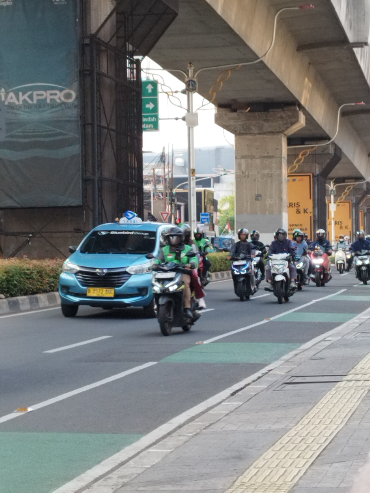 The image captures a street scene in South Jakarta, Indonesia, under an elevated structure, likely an overpass or highway. A light blue taxi with the "Bluebird Group" logo and a blue light on its roof is prominent in the foreground, driving on the right side of the road. The taxi has a yellow license plate with the number B 2573 BUC. In front of the taxi, a person on a motorcycle, wearing a green jacket and helmet, is also moving forward. A convoy of other motorcycles, each with a rider wearing a helmet, follows behind. The road has white lane markings and green painted sections. The elevated concrete structure above casts a shadow on the scene. On the left side of the image, a large banner with the word "AKPRO" is visible. Further down the road, on the right, there are yellow signs with text that includes "RIS & K." and "SPACE." The environment suggests a busy urban setting with ongoing traffic.