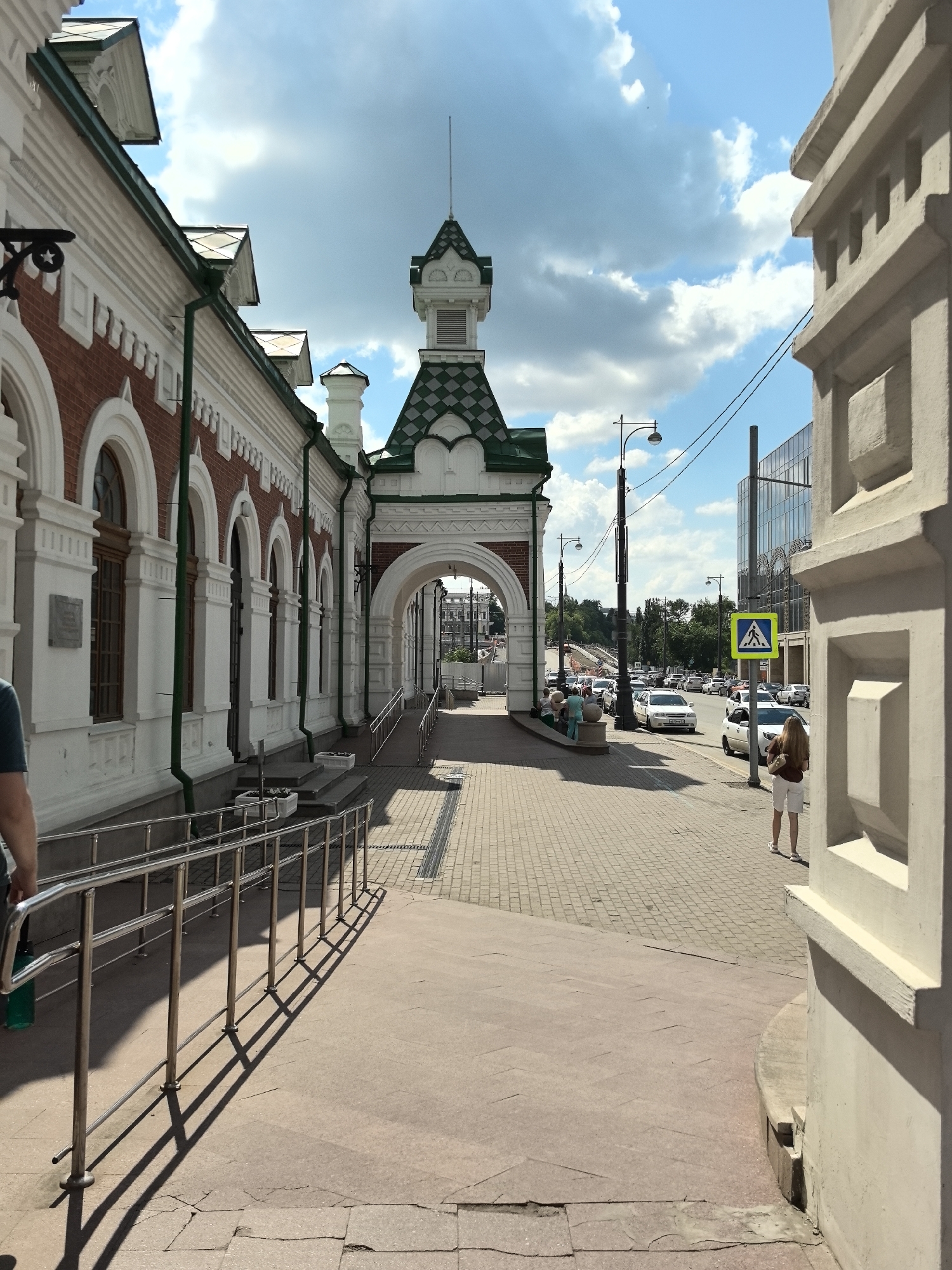 The image depicts a street scene in Perm, Russia, likely near a historical building with red brick and white architectural details, featuring an arched entryway with a green tiled roof. A paved sidewalk runs alongside the building, leading towards a street with vehicular traffic and pedestrians. In the foreground, a metal handrail runs along a ramp. A woman in white shorts and a brown shirt stands on the sidewalk near a crosswalk sign. The sky is blue with scattered white clouds.