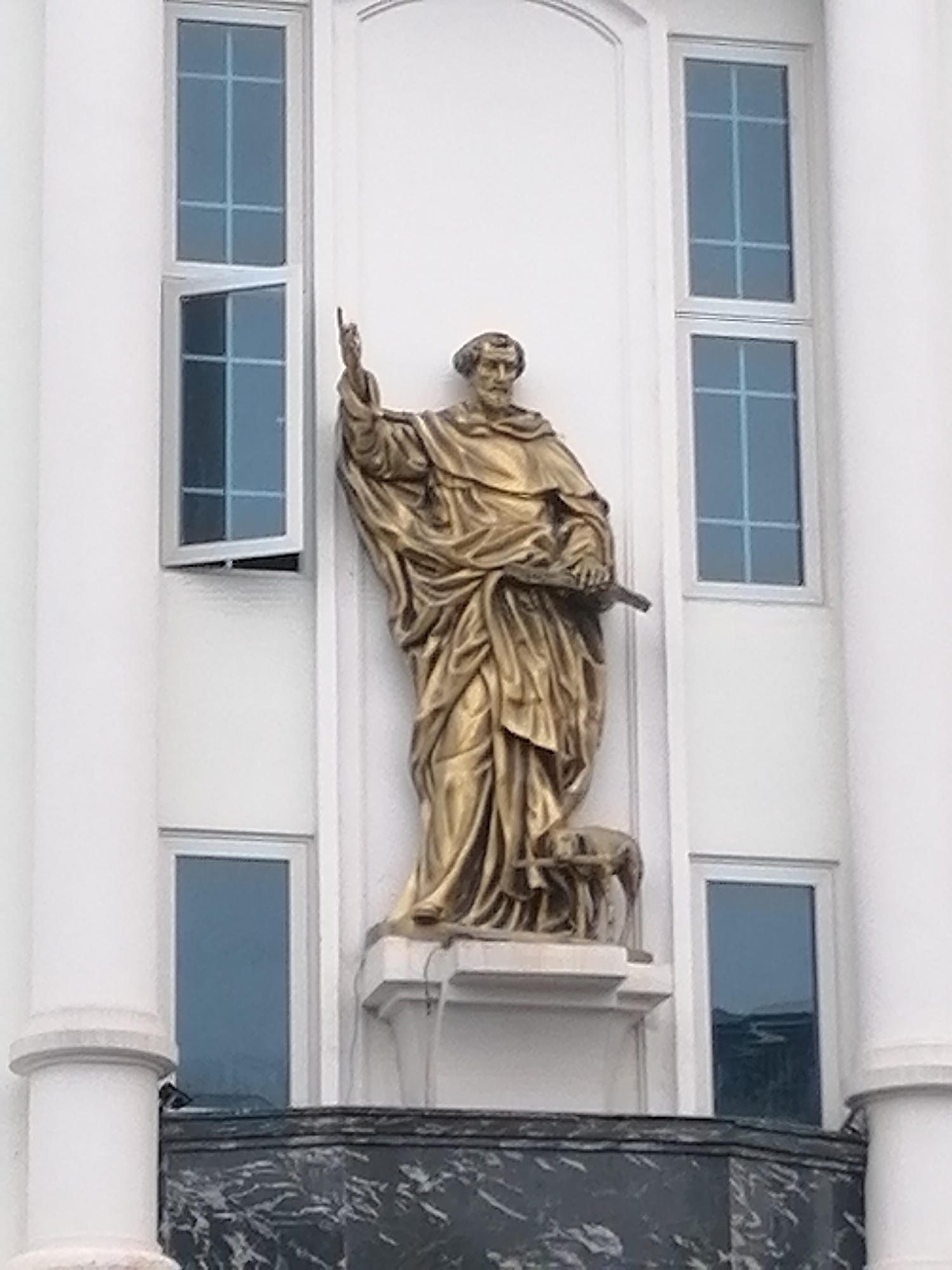 A medium shot shows a statue of a religious figure standing on a ledge in front of a white building. The statue is gold and depicts a man in robes with a raised right hand. He is holding a book or tablet in his left hand. A small animal, possibly a dog, is at his feet. The building behind the statue has white walls, pillars on either side, and several windows with blue reflective panes. One of the upper windows is partially open. The statue is located on a balcony with a gray, marble-like surface. The location is Da Lat, Vietnam.