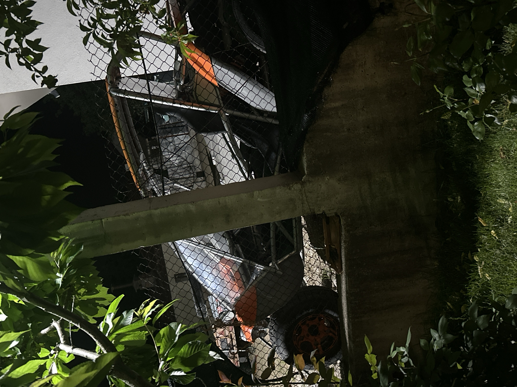 A nighttime view through foliage and a chain-link fence reveals a possible construction or demolition site. Structural beams are visible, and orange-colored machinery or equipment is partially obscured by the fence. The location appears to be outdoors, with vegetation present on either side. The time of day is dark, with artificial light possibly illuminating the scene.