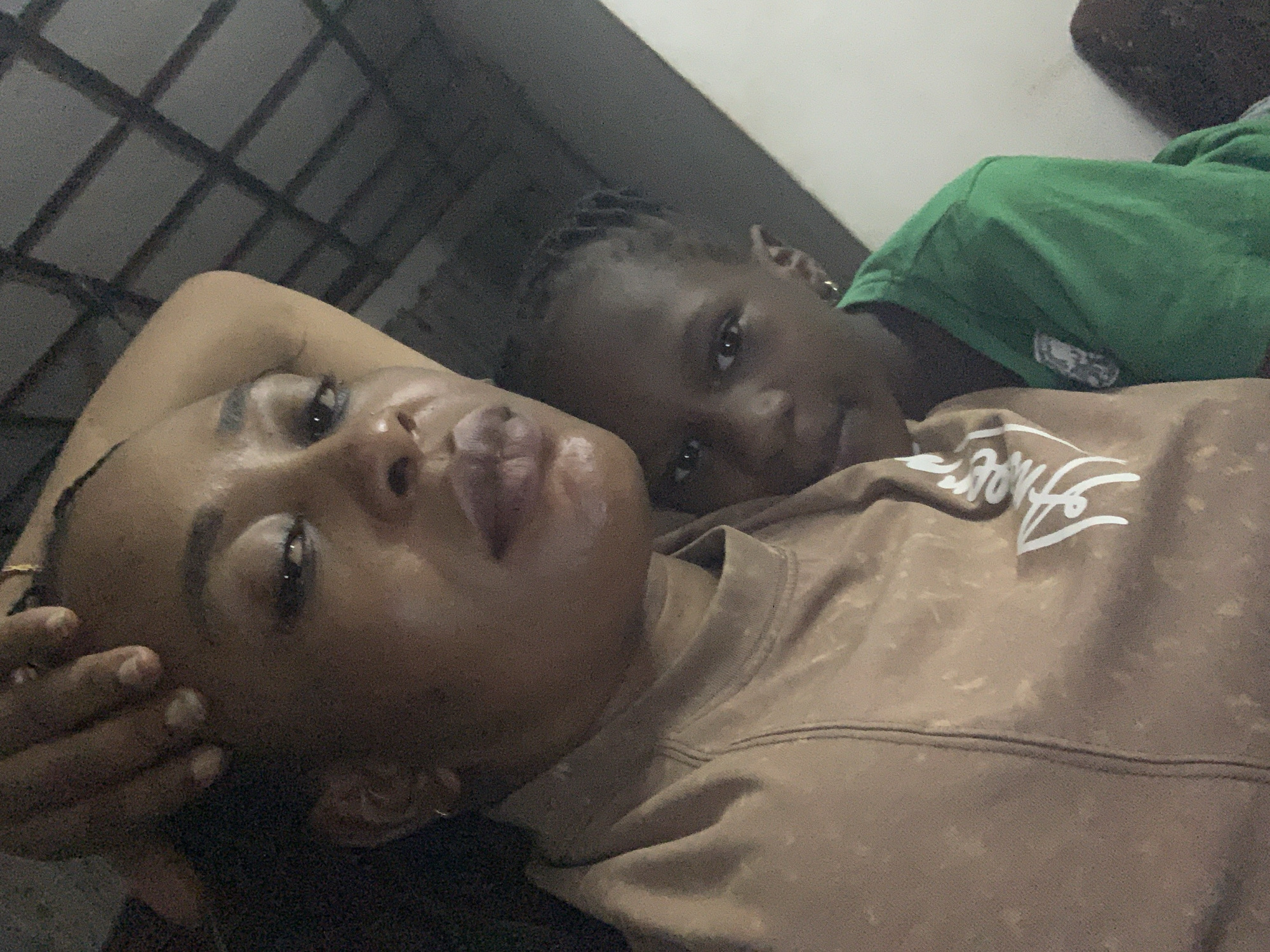 The image displays a close-up, dimly lit selfie of an adult Black woman and a young Black child, presumably taken indoors in Sabon Tasha, Nigeria.
The woman is positioned in the foreground, looking directly at the camera. Her dark hair is pulled back, and her right hand, with light-colored manicured nails, rests on her forehead. She wears a light brown or beige long-sleeved top featuring a subtle mottled pattern and a partial white, stylized logo visible on her lower front. Her lips are painted a dark berry shade, and a small white smudge is present on her chin. A thin hoop earring is visible in her left ear.
Behind and to the right of the woman, a child is partially visible, also looking directly at the camera. The child has dark braided hair and wears a vibrant green short-sleeved t-shirt with a small dark emblem on the chest. A small gold-colored hoop earring is visible in the child's right ear.
The background on the upper right shows a dark, textured surface with a distinct grid pattern, possibly a ceiling or wall. Below this, a lighter, plain wall section is visible. The left side of the background is dark and indistinct.