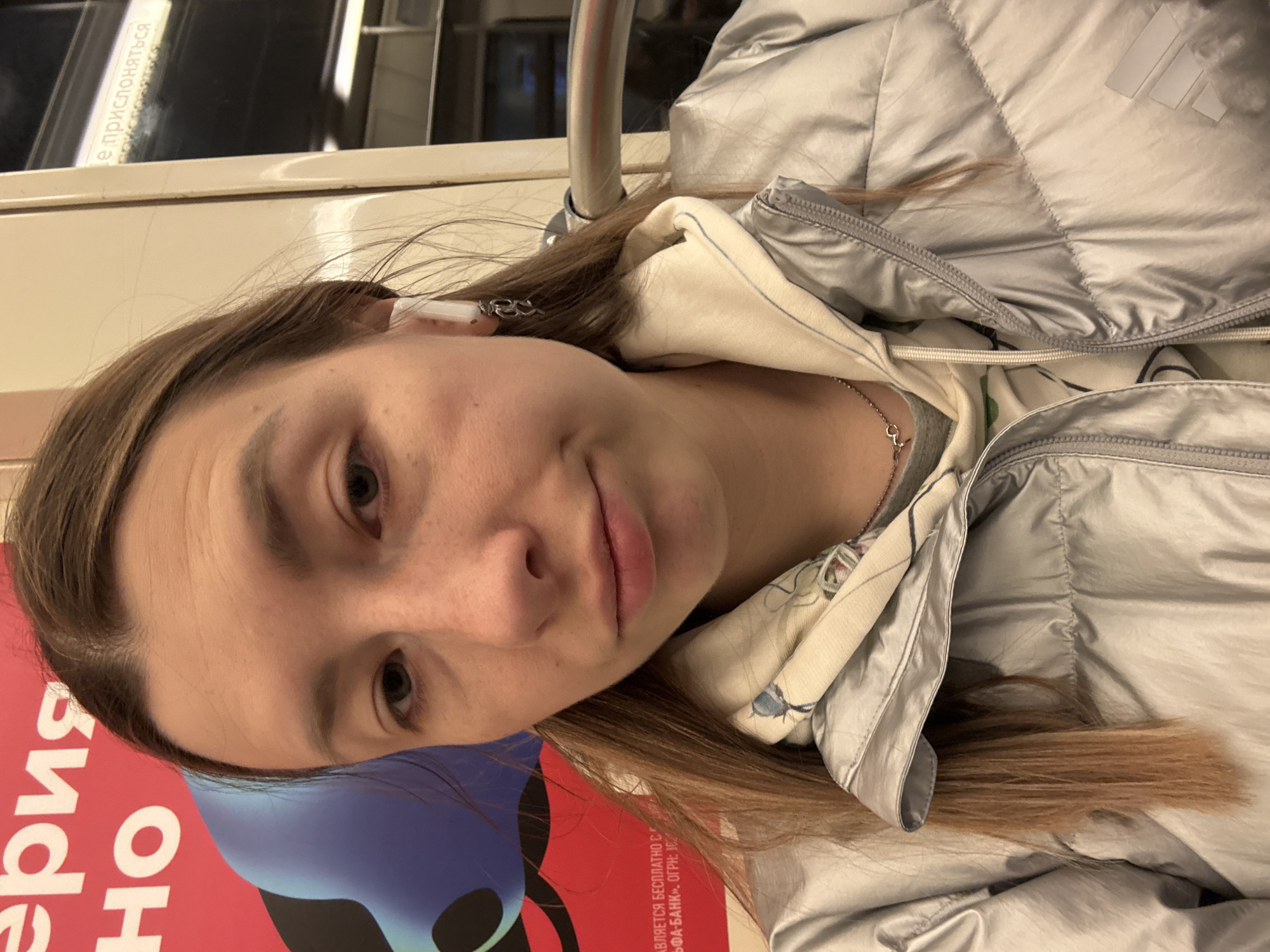 A portrait-style image captures a young woman inside what appears to be a public transport vehicle, likely a metro or subway in Moscow, Russia. The woman, who is the primary subject, has light brown, long hair, and fair skin. She wears a silver or light grey puffer jacket over a light-colored, possibly white or cream, hooded sweatshirt, which has subtle patterns. A silver chain necklace with a small pendant is visible around her neck, and she has a white earbud in her left ear. Her expression is neutral to slightly contemplative, with her lips softly closed. She is looking directly at the camera, suggesting a selfie or a close-up photo.
In the background, a metal pole or handle, typical of public transit, is visible. Above her head, a section of the vehicle's interior wall or ceiling is seen, bearing reflections. To the lower left of the frame, a vibrant red and blue advertisement or poster is partially visible.
Visible Russian text on a white sign in the upper background reads "Не прислоняться" (Do not lean), a common warning in public transport. On the advertisement below, fragmented text includes "является бесплатно с" (is free of charge with) and "Альфа-Банк" (Alfa-Bank), along with "ОГРН: 102", which is part of a legal registration number. These textual cues, combined with the Moscow context, confirm the setting as likely the Moscow Metro. The lighting is artificial, consistent with an indoor setting. The atmosphere appears calm and routine, reflecting a typical commute or travel moment.