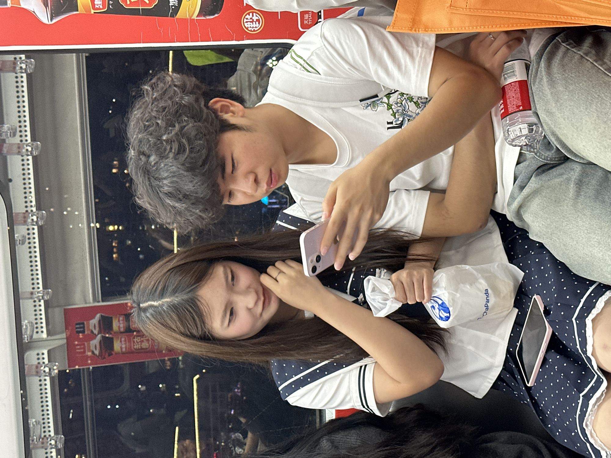 A young man and a young woman are seated, oriented toward each other and interacting with cell phones in what appears to be public transportation, likely a bus or light rail, based on the visible cityscape and adjacent vending machine. The man has curly dark hair, is wearing a white t-shirt, and is looking at his phone. The woman has long brown hair, is wearing a white and navy polka-dot top, and is also holding a cell phone as well as a white plastic bag, possibly from ChaPanda. They appear to be in Yuzhong District, China, indicated by the cityscape view through the window. A water bottle is also visible near the man, and there is an orange fabric object (possibly a bag) near him as well.