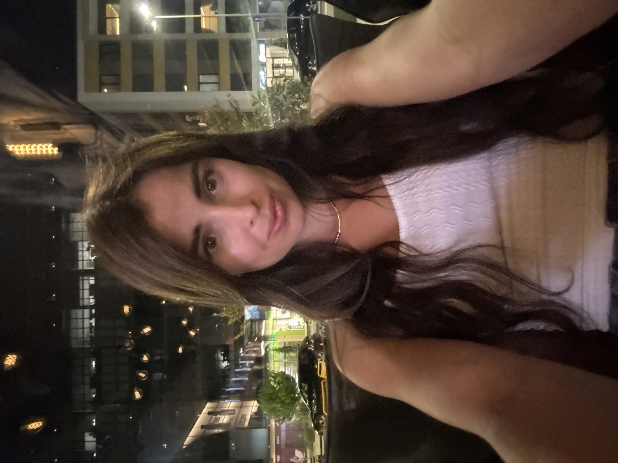 A young woman with long, dark, wavy hair is pictured in a close-up selfie. She is wearing a white, textured tank top and a delicate gold necklace. The background suggests an urban setting at night, with illuminated buildings and streetlights visible through what appears to be a large window or glass partition. Cars and greenery are also present in the background, indicating an outdoor or semi-outdoor area like a restaurant patio or a street-level view from an establishment. The lighting is dim, with some warm lights from the buildings and streetlamps creating reflections.