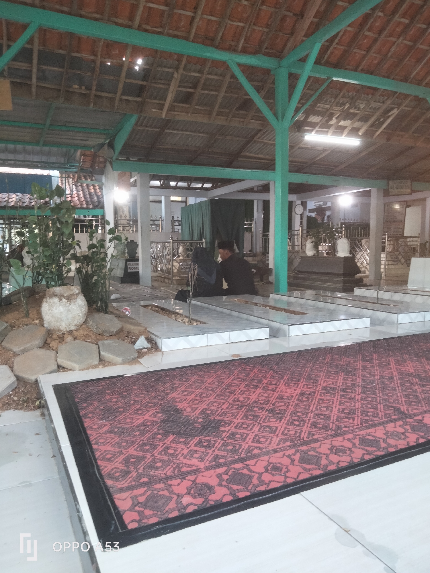 The image depicts an outdoor area with several graves. In the foreground, a large red rug with a geometric pattern is laid out on a tiled surface. Behind the rug, there are multiple graves, most of which appear to be simple stone slabs. To the left, there is a small garden area with plants and stones. In the background, a covered structure with a tiled roof supported by green pillars is visible. The structure appears to be a mausoleum or a shelter over graves. There are at least two individuals present, one wearing dark clothing and a cap, and another person dressed in dark clothing. They are near one of the graves, suggesting a memorial or prayer setting. The overall environment appears to be a cemetery or a sacred burial ground.