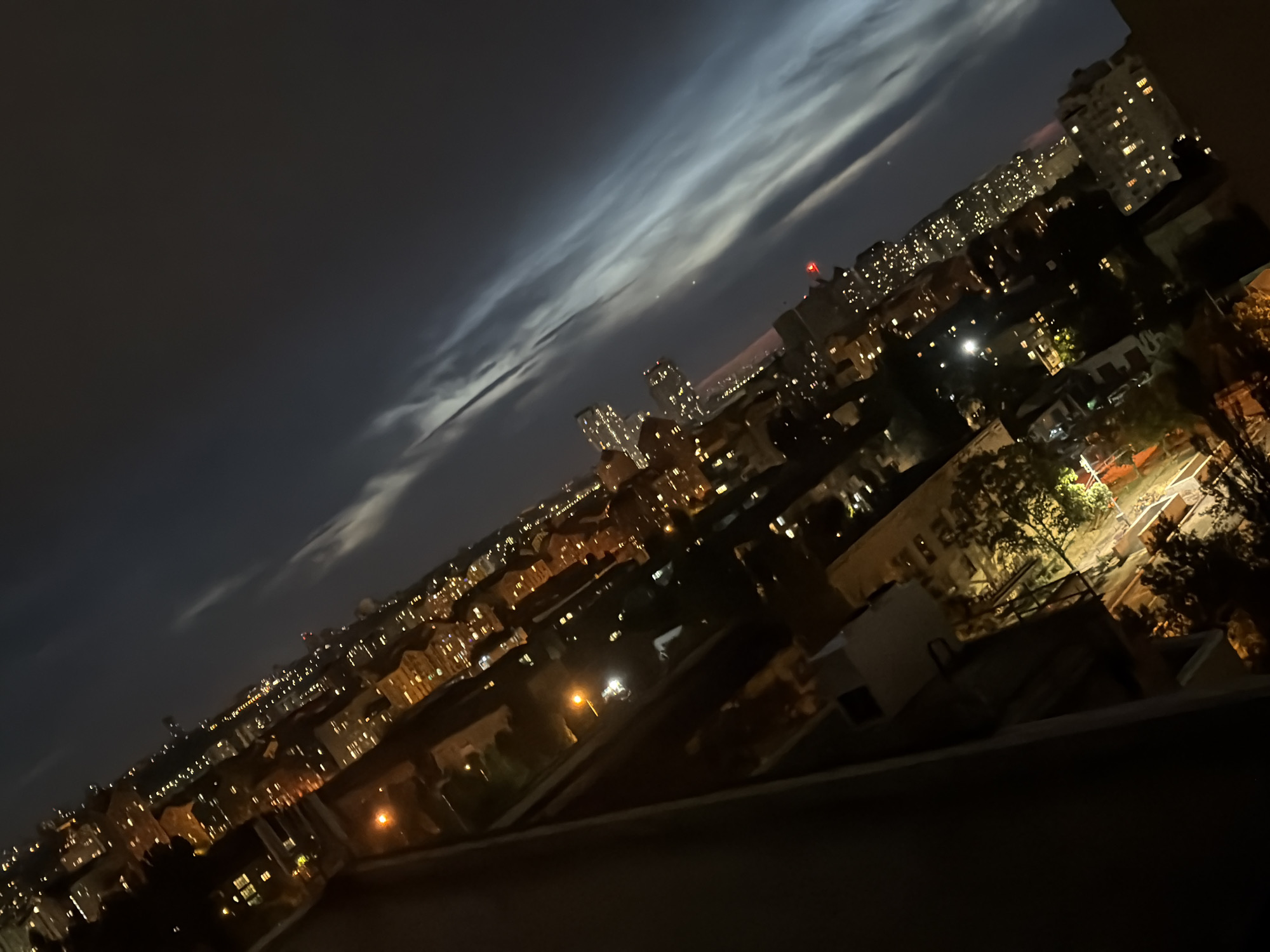 An angled, nighttime, aerial view of a city skyline, likely Kyiv, Ukraine, is depicted. The scene is dominated by the warm glow of numerous building lights against the dark sky, suggesting an urban environment with a dense population. Streaky, luminous clouds stretch across the upper portion of the frame, illuminated by ambient city light, creating a dynamic contrast with the deep blue-black of the night sky. In the foreground, a dark, angled structure, possibly a building or balcony railing, partially obscures the view. The city lights create a textured pattern across the buildings, with some taller structures and their individual windows clearly discernible. The overall impression is one of a vibrant, active city at night, with the natural atmospheric phenomenon of the illuminated clouds adding a surreal quality to the urban landscape.