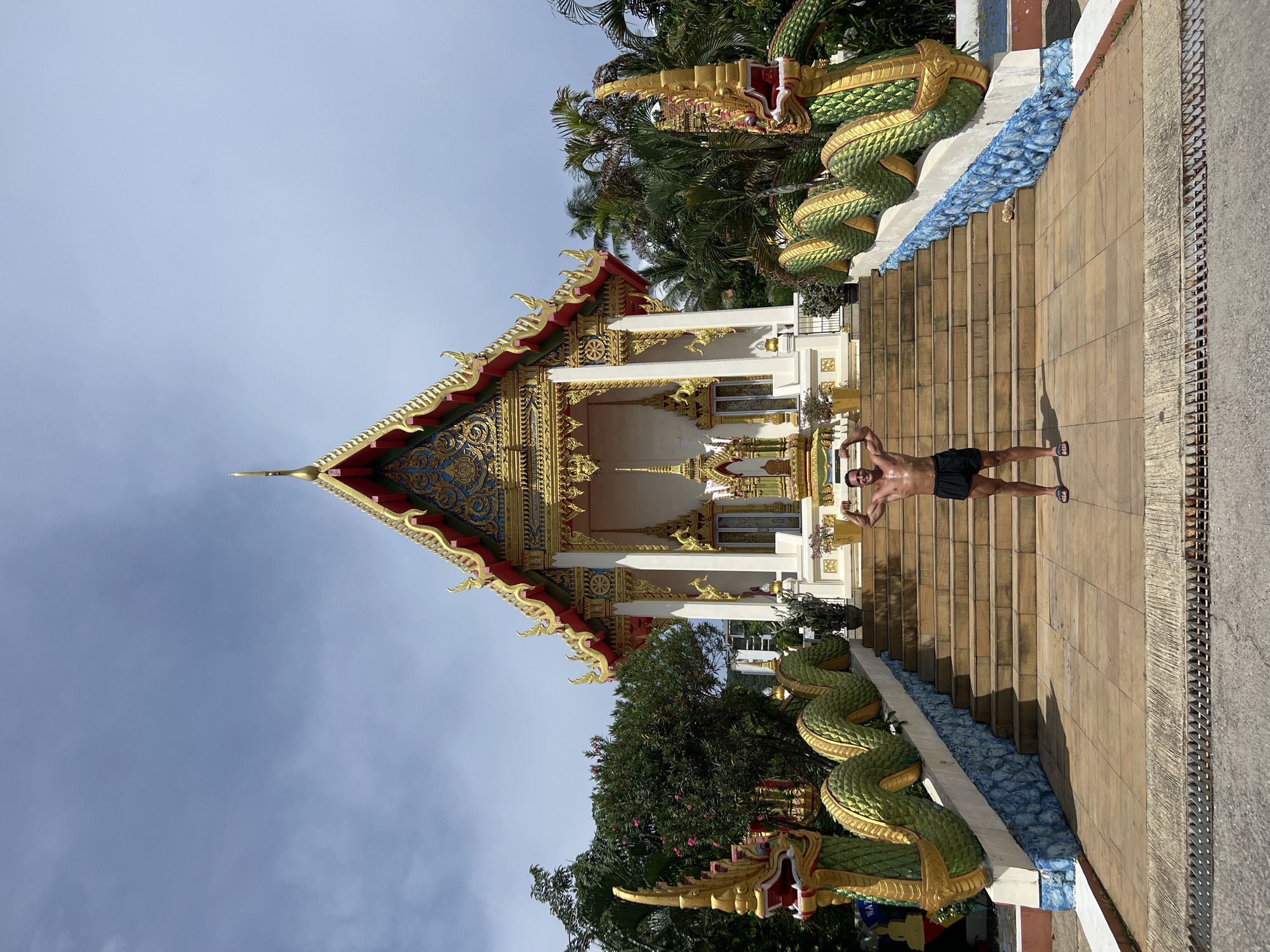 A shirtless man with dark hair and a muscular build, wearing black shorts and flip-flops, poses with his arms flexed on the stairs of a temple. He is centered in the frame, facing the viewer. The temple, located in Karon, Thailand, features a white facade with ornate gold and red trim on its roof. Two large, green, dragon-like statues with gold accents flank the entrance stairs, which are lined with blue mosaic tiles. Lush green trees and palm trees surround the temple, and the sky is partly cloudy.