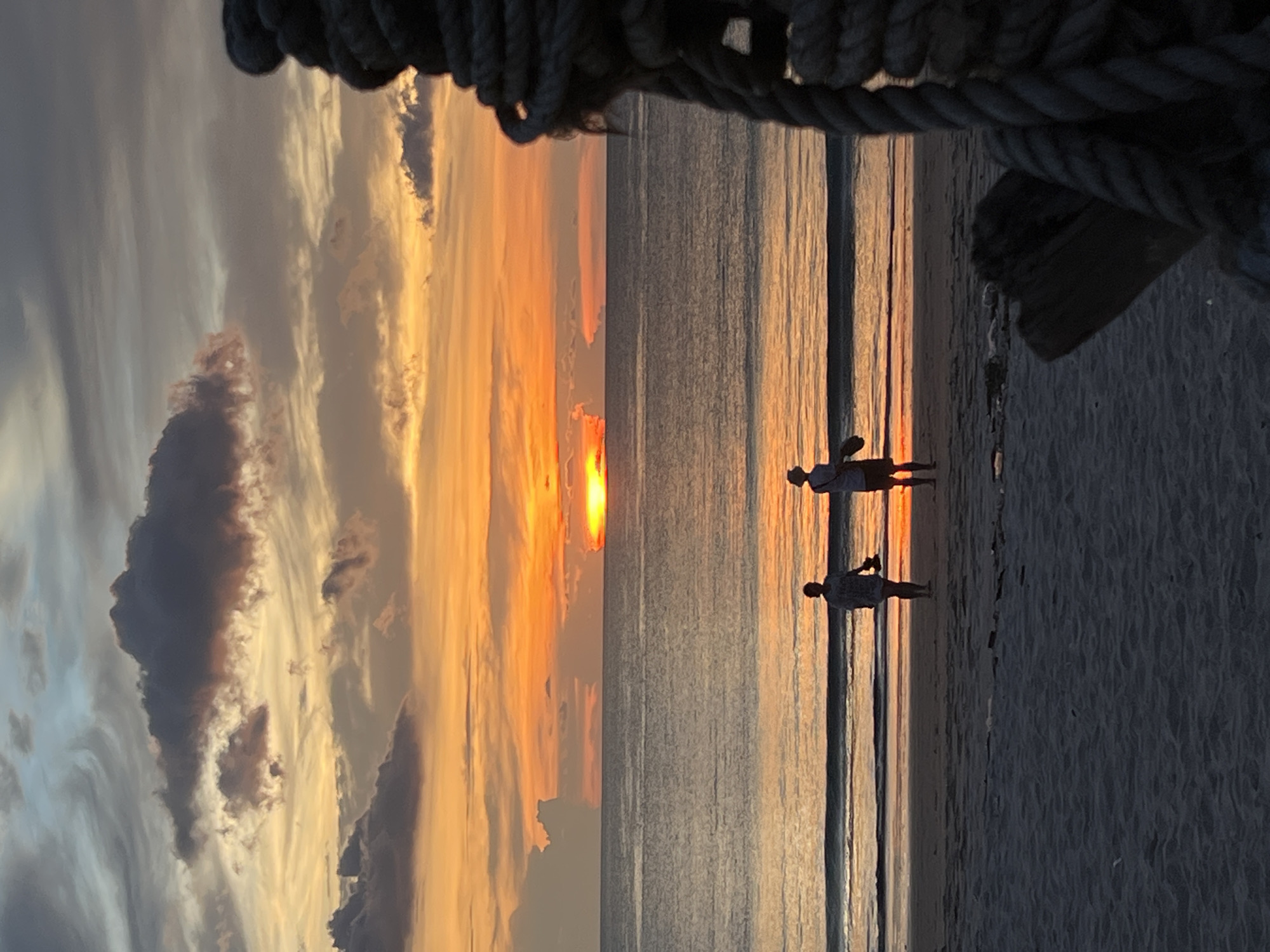 Two figures, presumably people, are standing on a beach at sunset. The location appears to be in an unknown city. The sky is filled with clouds, illuminated by the setting sun in hues of orange and yellow. The sea reflects the colors of the sunset. A rope, likely part of a boat or structure, is visible at the top of the image.