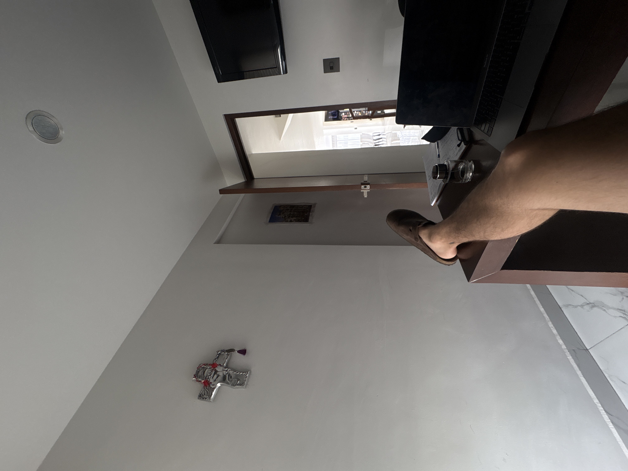 The image depicts an interior space, likely a room in a residence, featuring a desk, a laptop, and a person's leg and foot in a brown loafer. The desk is positioned near a doorway that leads to another room, visible in the background. A television is mounted on the wall. Additionally, a decorative cross and a framed image hang on the wall. The background suggests the location could be in San Bernardino Tlaxcalancingo, Mexico, although there are no specific landmarks or identifiers visible in the image to confirm this location.