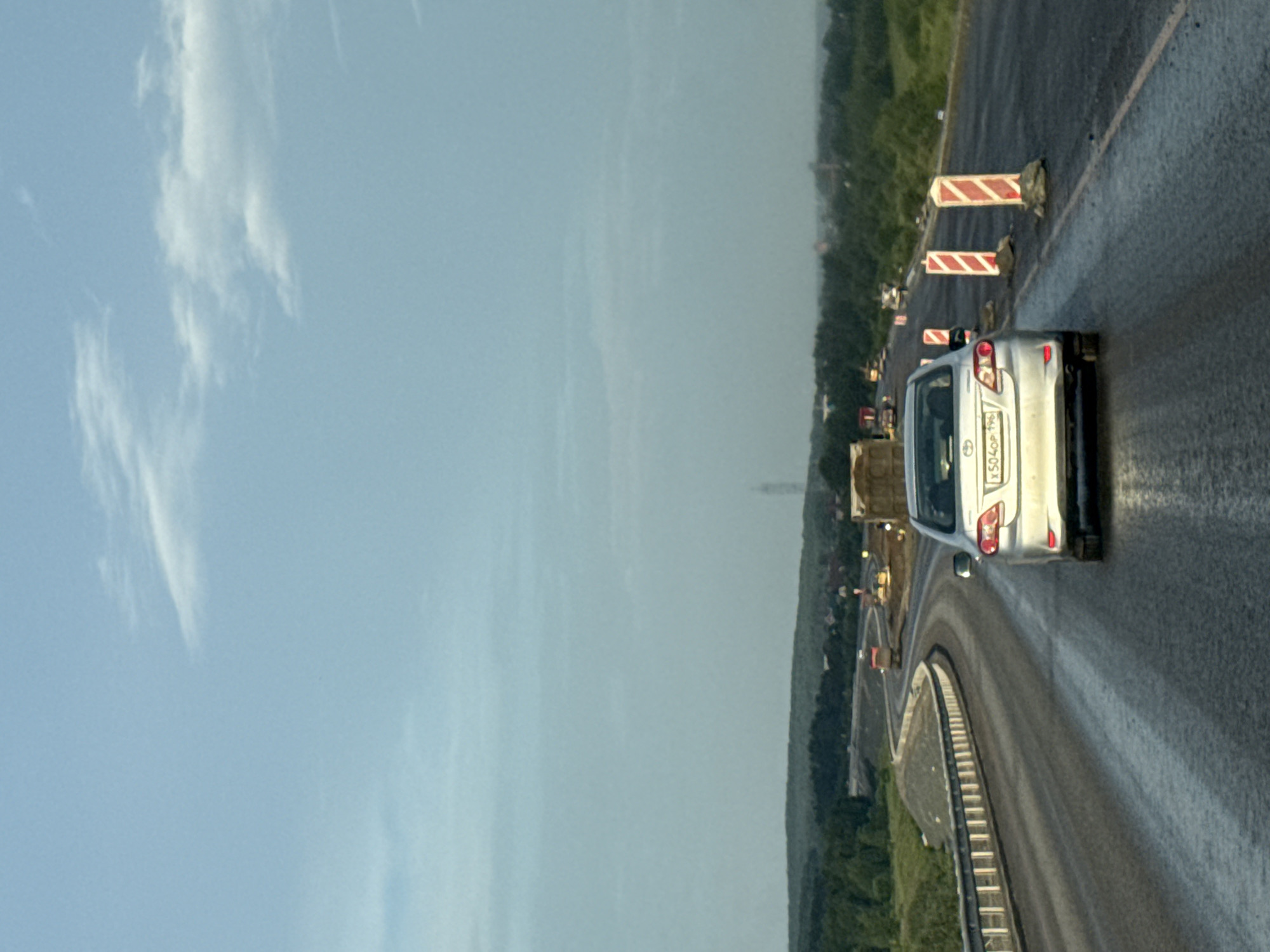 A silver Toyota Corolla is traveling on a wet highway. The car has a license plate that reads "X504OP 196". The highway appears to be undergoing construction, with orange and white traffic cones and barriers visible. In the distance, there are trees and buildings, and a tall structure resembling a communication tower. The sky is overcast with some clouds. The image is taken from a slightly elevated perspective, looking forward from behind the vehicle. The location is identified as the city of Boyarka, Russia.