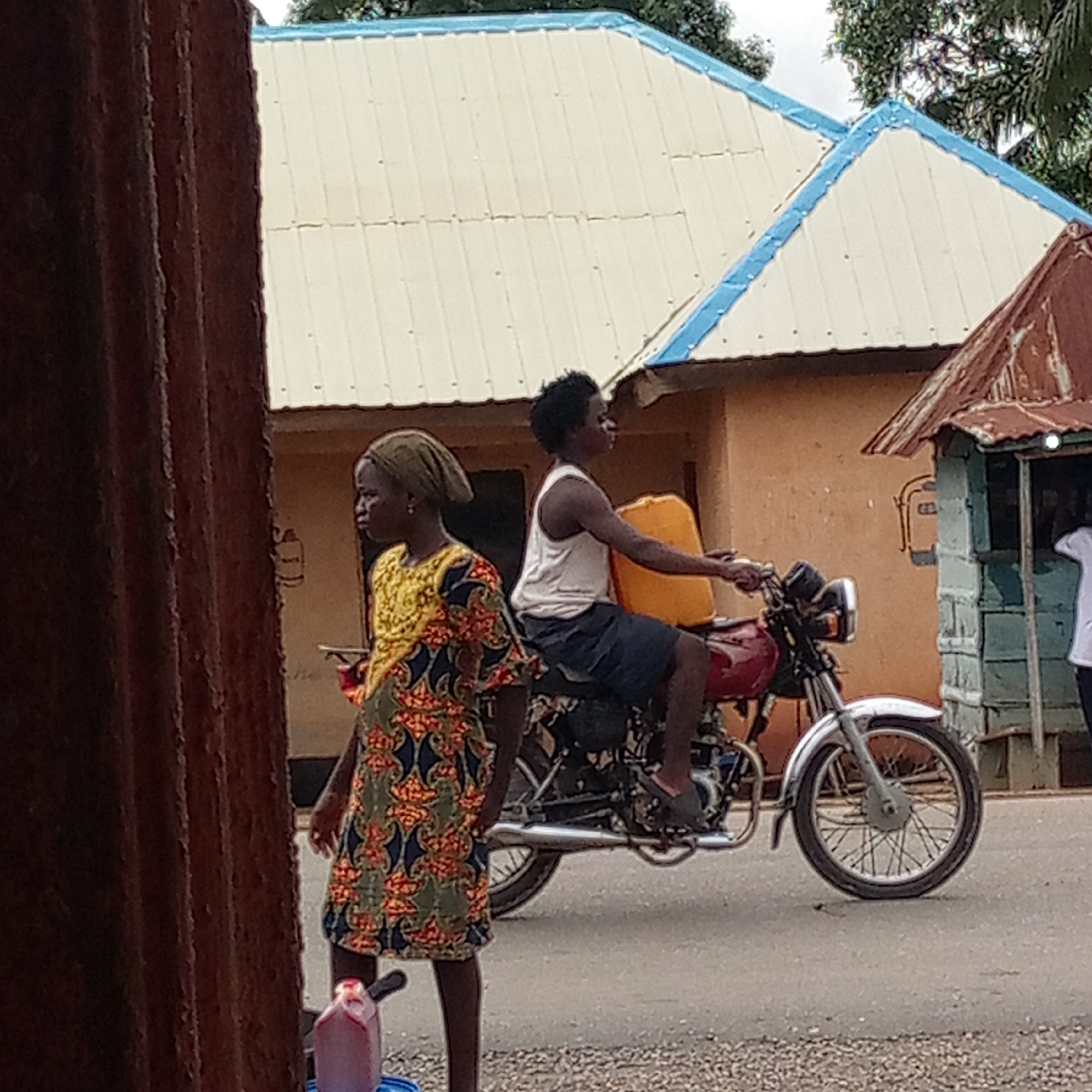 In Mararaba, Nigeria, a woman in a green and yellow patterned dress and headscarf stands beside a red container on the left side of a street. A man rides a red motorcycle along the same street, carrying a yellow jerrycan strapped behind him. Behind them, there's a house with a white roof edged in blue. To the right, there's a small structure that is green and blue. The street is paved, and a weathered corrugated metal sheet is visible on the extreme left of the frame.