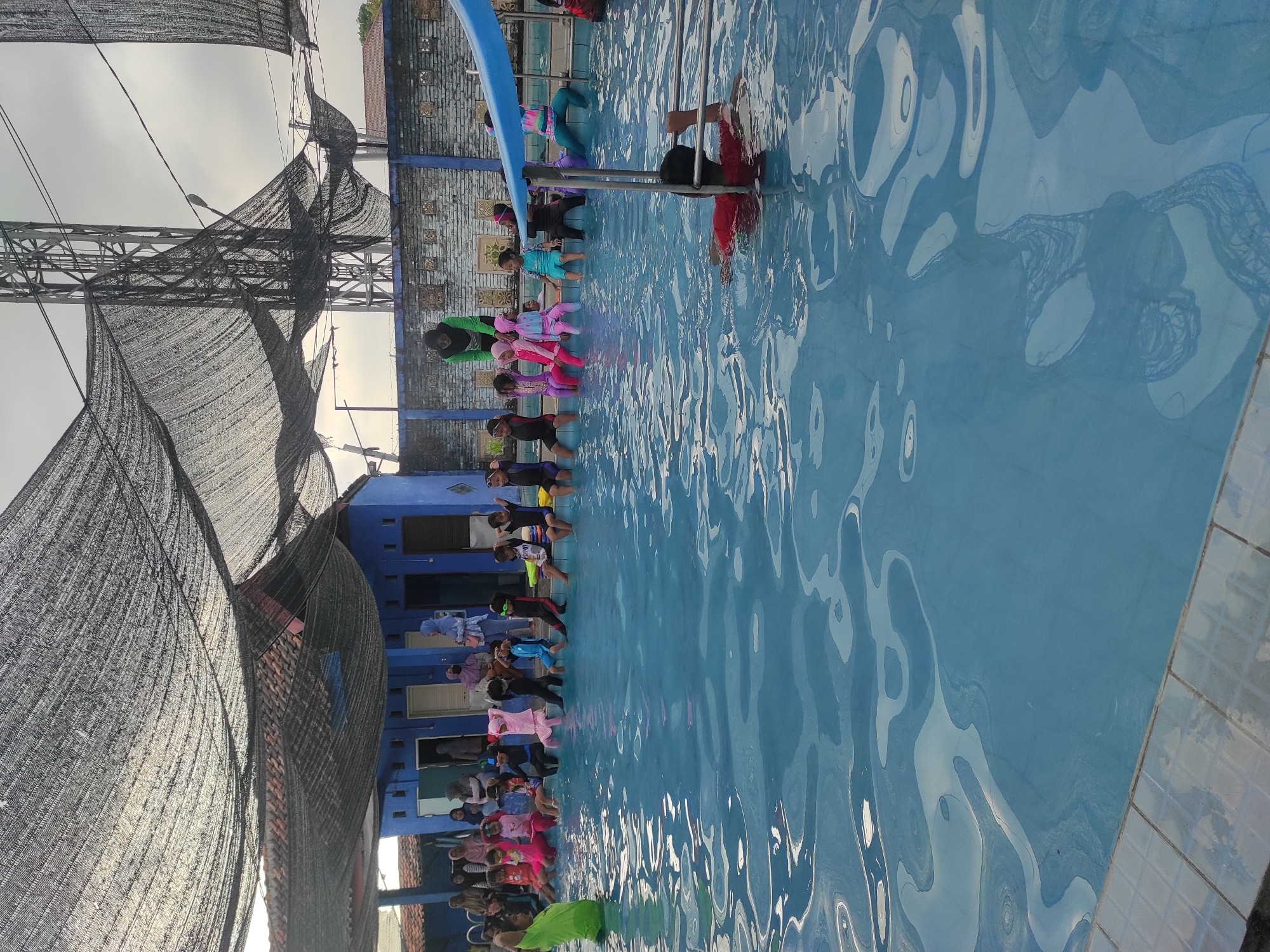 The image depicts an outdoor swimming pool area in Pleret, Indonesia, under a large, dark mesh sunshade. A group of children, dressed in various swimsuits and some wearing headscarves, are seated on the edge of the pool, with their legs dangling in the water. An instructor wearing a bright green shirt is visible on the left, facing the children and appearing to be giving instructions. To the right, a person in a red shirt is partially submerged in the pool, seemingly swimming. In the background, a blue building with several doorways is visible, and beyond that, a brick wall and a metal tower structure. The pool water is clear blue, reflecting the overcast sky and the overhead sunshade.