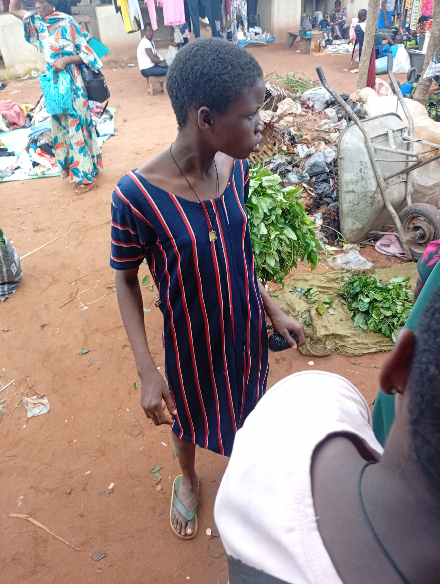 The image depicts a busy outdoor market scene in Nkpor, Nigeria. In the foreground, a young Black female stands with her head turned to the side. She is wearing a dark blue dress with vertical red and white stripes and a necklace with a yellow pendant. She has dark, short hair and is barefoot, wearing a green flip-flop on her left foot. Her right hand is extended slightly downwards, and she appears to be holding something black.
Behind her, a woman in a colorful dress with a floral pattern and a blue scarf is visible. She is carrying a blue bag in her left hand and a black bag slung over her shoulder. Other people and market stalls are scattered throughout the background. The ground is covered in reddish-brown soil and debris. A wheelbarrow is visible on the right side of the image, filled with what appears to be trash and bundles of green leaves. In the middle ground, there are more bundles of green produce laid out on a cloth. The general atmosphere suggests a lively and crowded marketplace.