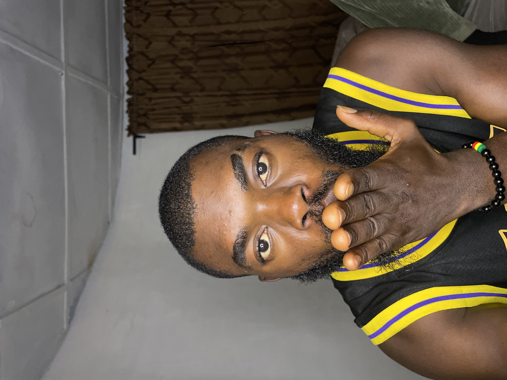 The media file shows a close-up, vertically oriented shot of a Black man's face, with his hand covering his mouth. He has a beard and is wearing a black jersey with yellow and purple trim. A beaded bracelet with red, green, and black beads is visible on his wrist. The background is indistinct, with a textured pattern visible above the man's shoulder and a plain wall on the left. The lighting suggests an indoor setting, possibly at night or in a dimly lit room. The man's eyes are open and looking directly at the camera, conveying a neutral or contemplative expression. The image is cropped to focus on the man's face and upper body. There is no visible text in the media file. The provided location context is Jos, Nigeria.