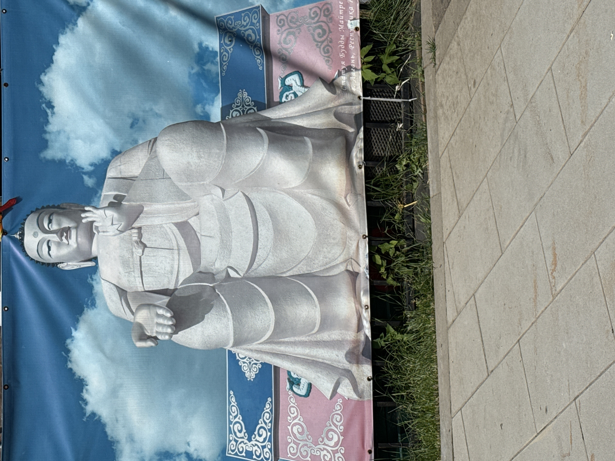 A large banner depicting a white Buddha statue against a blue sky with clouds is displayed outdoors. The banner is attached to a fence or barrier, with green grass and a paved walkway in the foreground. Text is visible at the bottom right of the banner, partially obscured by grass, which appears to be in Cyrillic script. The scene suggests a public space, possibly a park or street, in Moscow, Russia. The banner itself is the primary subject, with the Buddha statue rendered in a seated position with one hand raised. The surrounding environment is secondary, providing context for the banner's display.