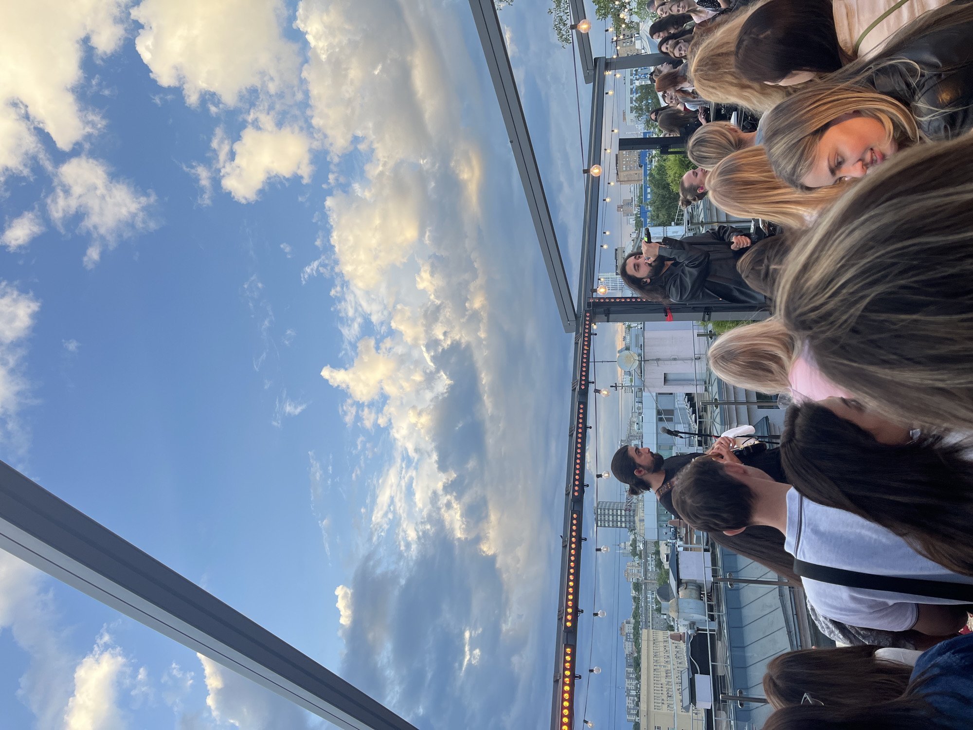 Here is a forensic description of the image.
The image is taken outdoors, seemingly on a rooftop venue in an urban environment. The sky is visible and filled with clouds. The sky is reflected in a glass rooftop, adding a layer of visual complexity. There are multiple people present, most of them appearing to be an audience. There is a musician who is performing. There is a lighting structure that holds rows of lights. Buildings are visible in the background, indicating an urban environment.
