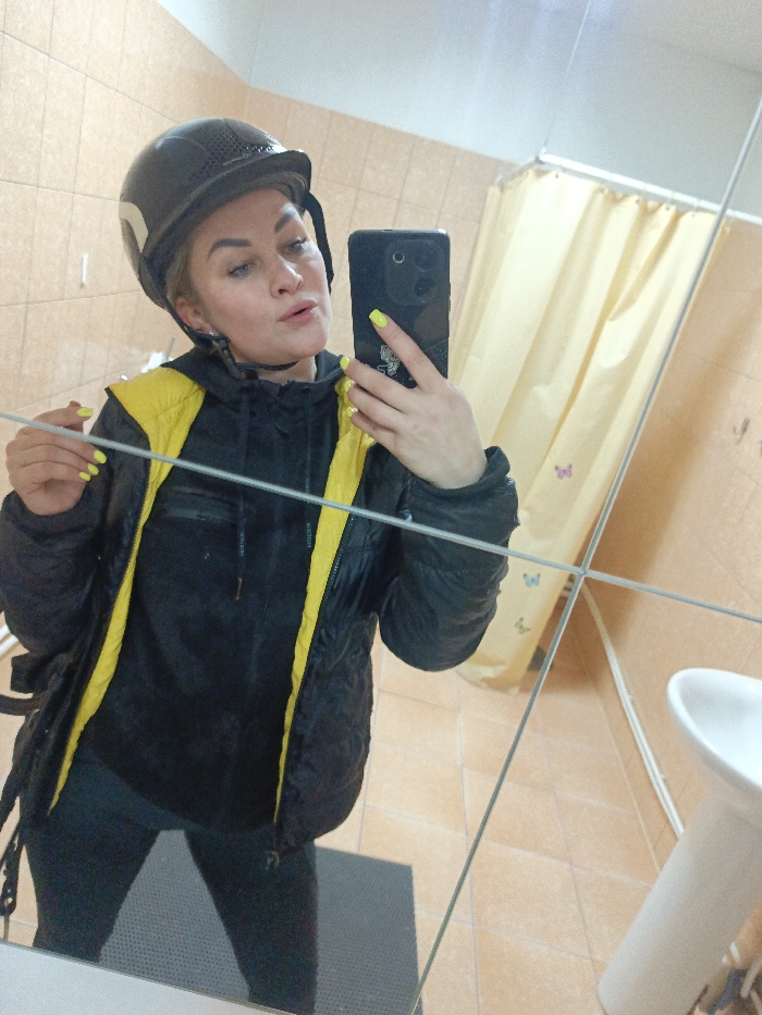 The scene occurs indoors, in what appears to be a tiled bathroom or changing room, identified as being in the city of Gorka, Russia. A female individual, approximately from the mid-thigh up, is centered in the frame, captured in a mirror selfie. She wears a dark-colored riding helmet with a white stripe on the left side. Her facial features include defined eyebrows, full lips, and light-colored eyes looking directly at the camera. She is dressed in a black puffer jacket with a bright yellow lining and internal trim, worn open over a dark long-sleeved top or hoodie. Below the jacket, dark-colored leggings or trousers are visible. Her left hand holds a dark-colored smartphone, featuring multiple camera lenses, positioned to capture the reflection. Her fingernails are painted in a bright yellow/neon green shade, with some white accents on certain nails. Her right hand is lowered, positioned near her chest. The background shows light brown square tiles on the walls and floor. To the right, a yellow shower curtain, adorned with multiple small, multi-colored butterfly decals, is partially visible. A white pedestal sink is also present on the right side of the frame. A dark grey or black rectangular mat is visible on the tiled floor in the lower central area. The mirror appears to be composed of multiple panels, indicated by intersecting lines creating a grid-like reflection.