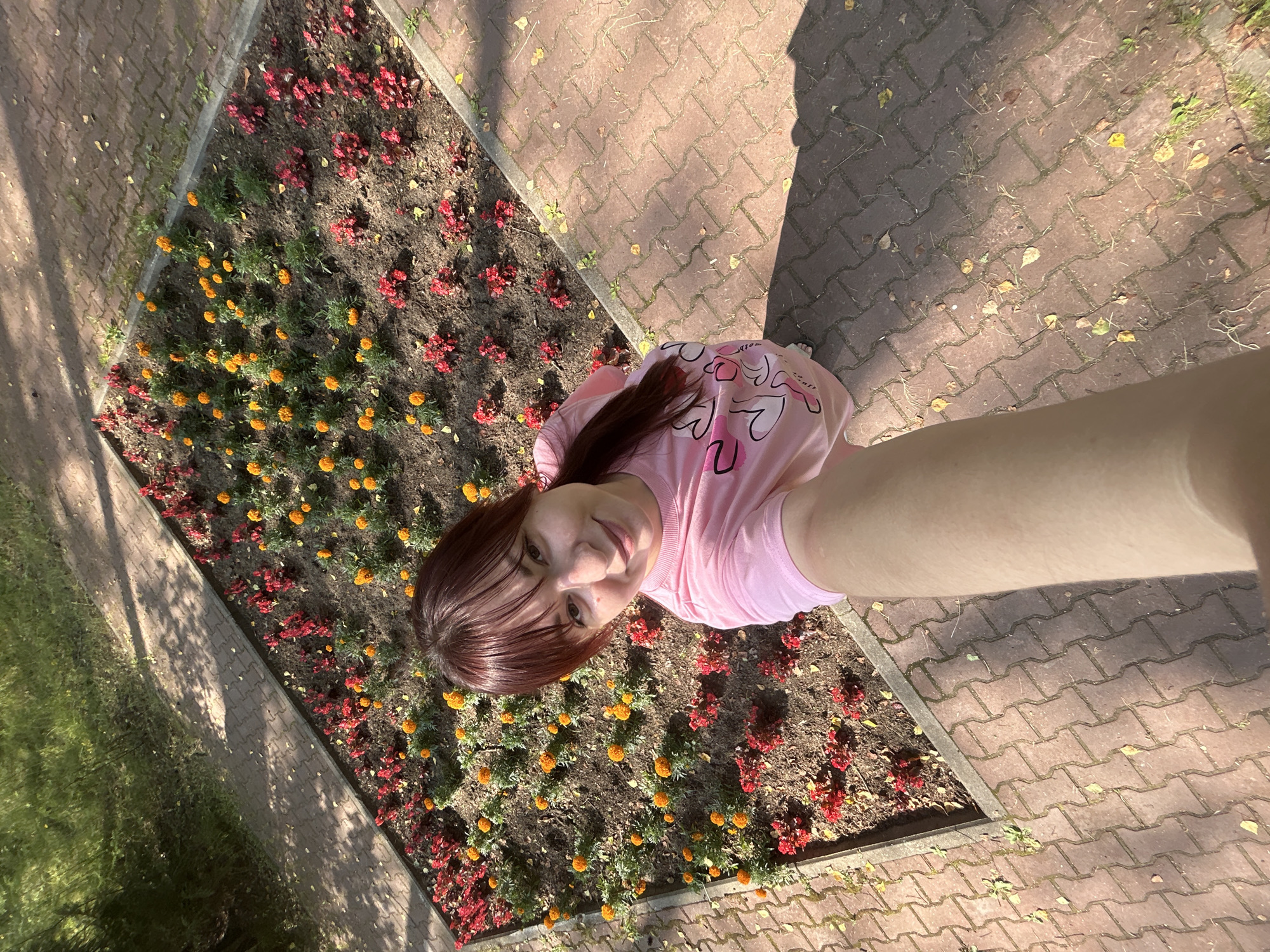 In Krasnovishersk, Russia, on a paved walkway, a young woman with reddish-brown hair takes a selfie. She is wearing a pink t-shirt adorned with white hearts and is positioned in front of a flowerbed. The flowerbed is rectangular and contains a mix of red and orange marigolds. The walkway is made of red-brown interlocking pavers, and scattered leaves indicate an autumn setting. Sunlight casts shadows across the scene. The woman's arm extends towards the camera, capturing her face and upper body.