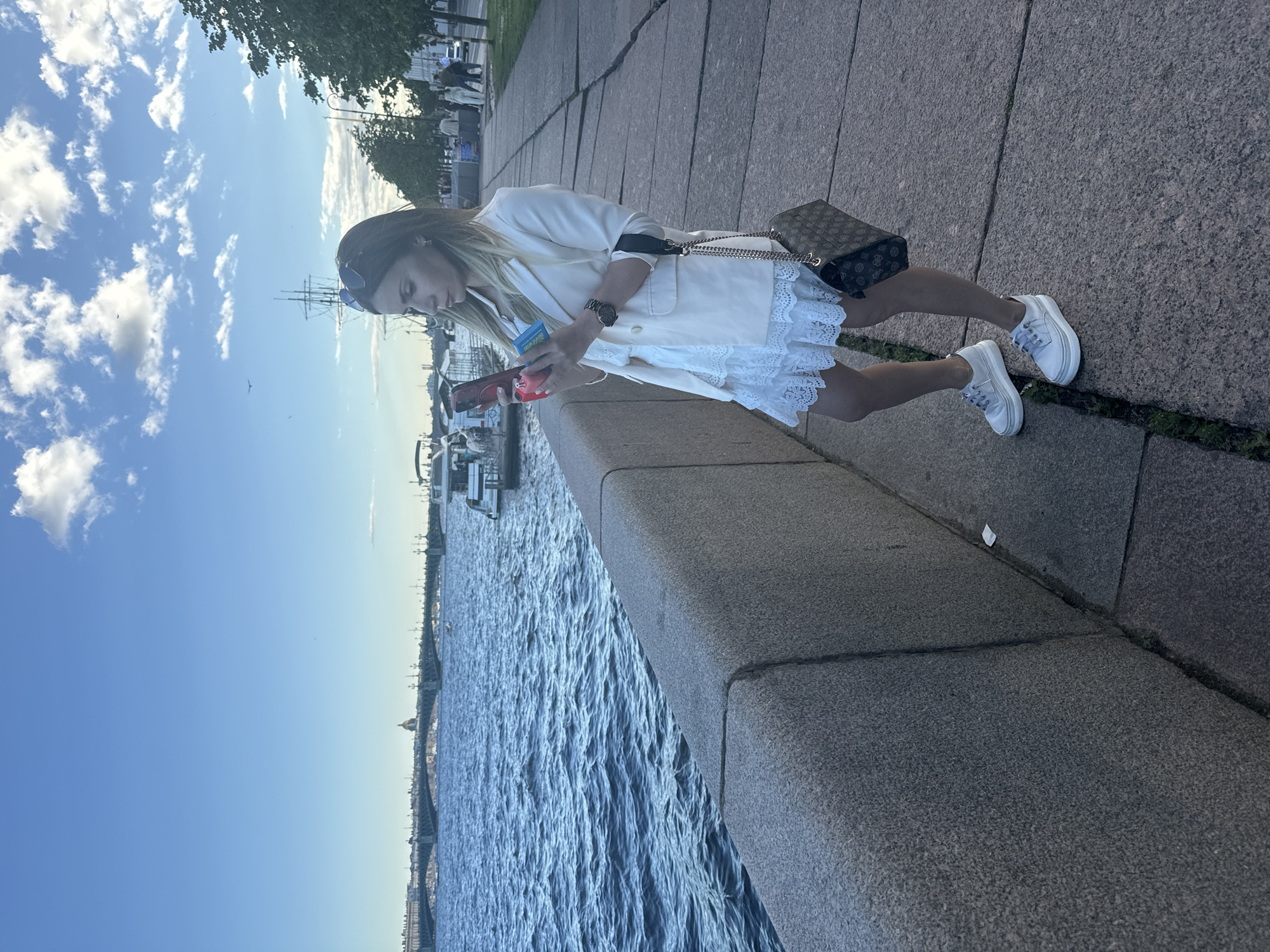 A woman stands on a stone promenade next to a body of water in Saint Petersburg, Russia. She is wearing a white lace dress, a cream-colored blazer, and white sneakers. She is holding two phones and looking at one of them. A brown patterned purse with a gold chain strap hangs from her shoulder. In the background, a bridge spans the water, with buildings and a large dome visible on the far bank. Several boats are docked or moving in the water. The sky is blue with scattered white clouds, and trees line the promenade to the right. The scene appears to be set in the late afternoon or early evening.