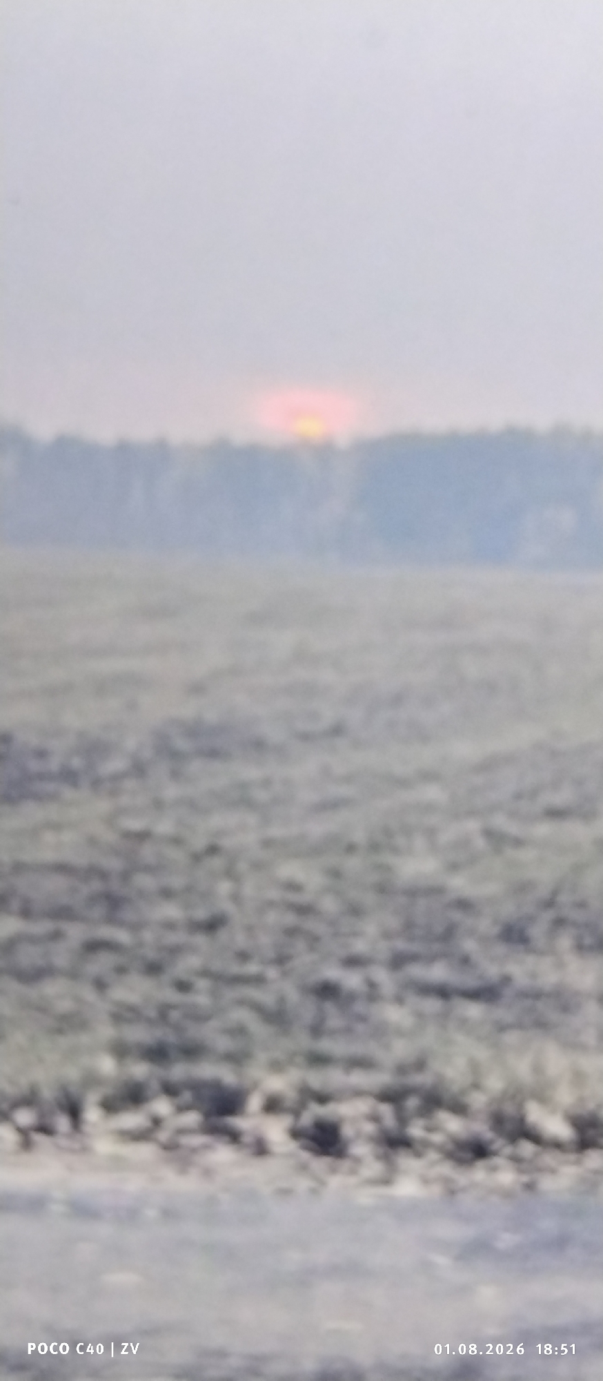 The image depicts a landscape scene at dusk, with a blurred sunset visible above a treeline in the distance. The foreground consists of a textured, grayish surface that appears to be either frozen ground or shallow water. The lighting suggests the time is around sunset, with a soft, diffused light illuminating the sky. The bottom left corner of the image shows the text "POCO C40 | ZV," indicating the device used for capture. The bottom right corner displays the date and time "01.08.2026 18:51." The overall scene is hazy and out of focus, making it difficult to discern specific details of the environment. Given the location context of Лубянки, Russia, this could be a view of a rural or natural area outside the city, possibly near a body of water or open field.
