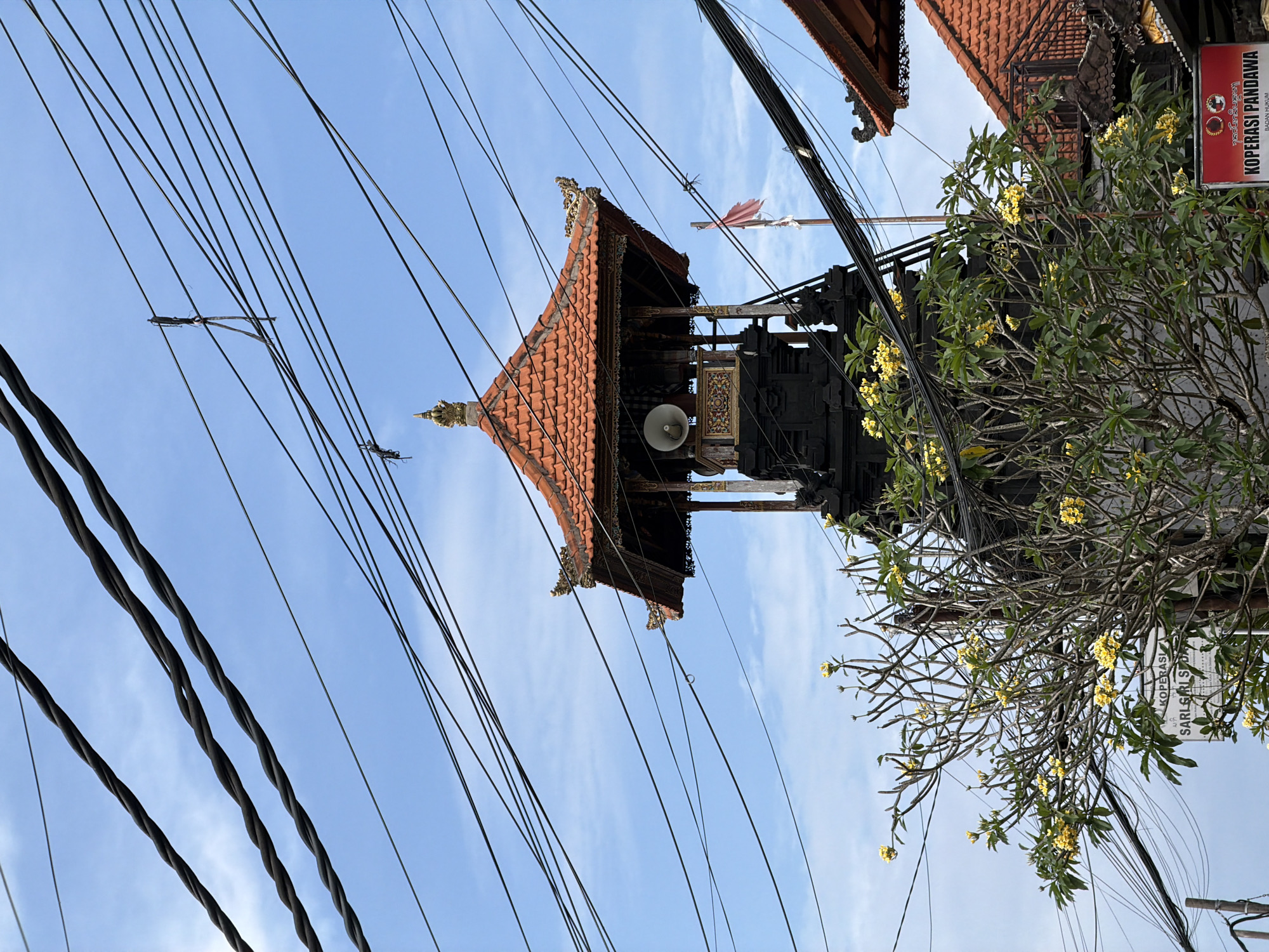 In Kuta Selatan, Indonesia, a tall, dark, stone structure with a red-tiled roof stands partially obscured by an abundance of electrical wires crisscrossing the sky. This structure appears to be a tower or bell tower, with a speaker visible at its apex. A small, tattered red and white flag flutters on a pole to the right of the tower. In the foreground, a tree with sparse green leaves and numerous yellow flowers blooms, its branches reaching towards the tower. The background consists of a bright blue sky with scattered white clouds. To the lower right, a portion of another red-tiled roof is visible, suggesting a cluster of buildings. A red sign with white text that reads "KOPERASI PANDAWA" is also present in the lower right corner, indicating a cooperative business.