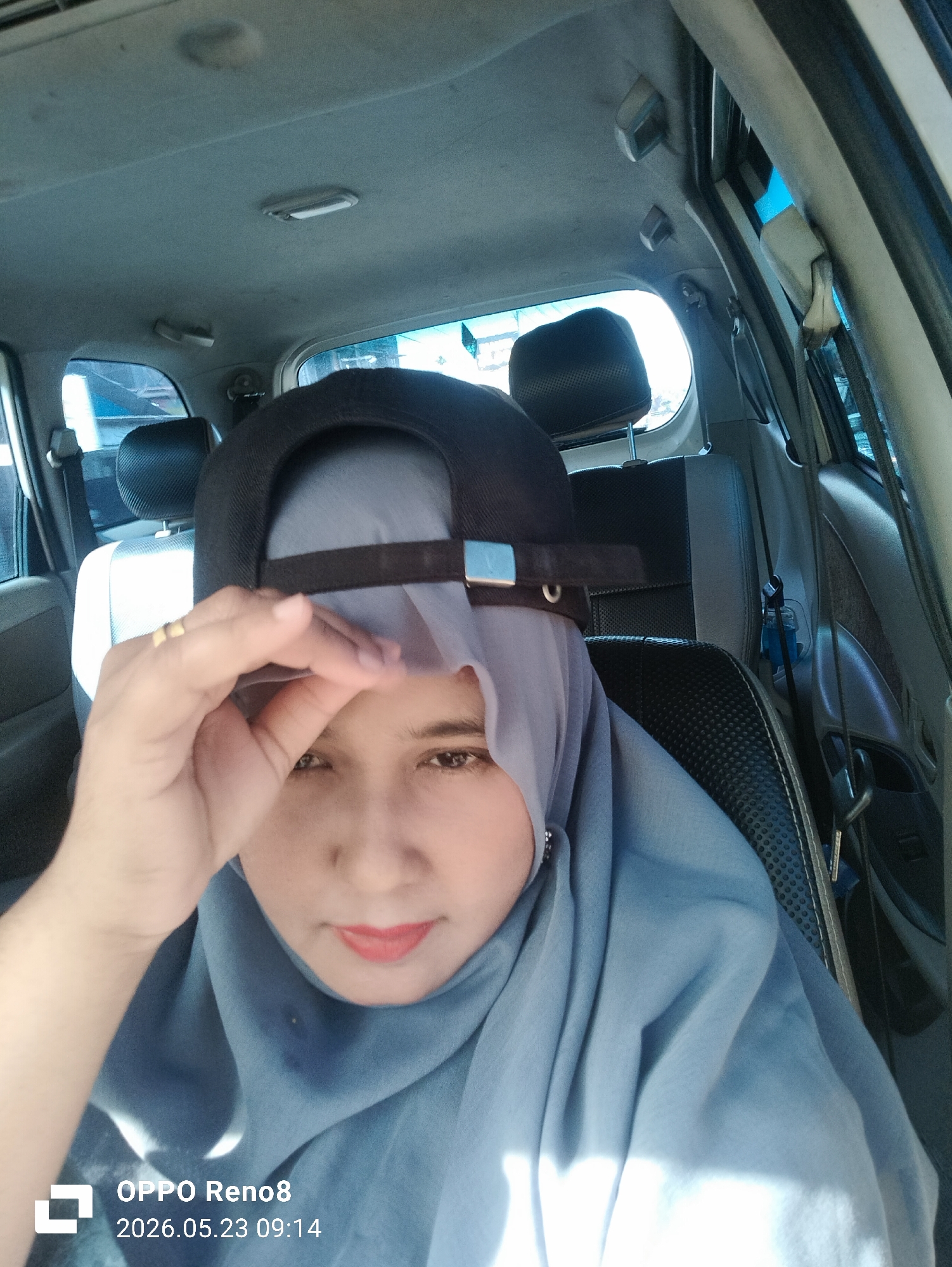 A woman wearing a light blue hijab and a black baseball cap is seated in the driver's seat of a car. Her right hand is raised to the brim of the cap, obscuring part of her face. The car's interior is visible, including the front passenger seat and headrest, along with the dashboard and window. The location is suggested to be in Teungoh Baro, Indonesia. The camera used is an OPPO Reno8, and the timestamp indicates the photo was taken on May 23, 2026, at 09:14.