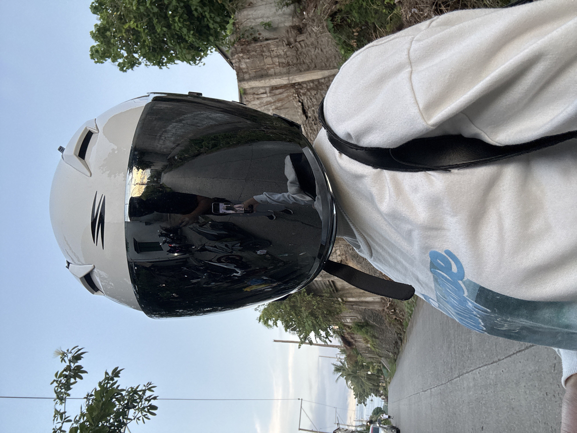 A first-person perspective shot shows a person wearing a white helmet with a reflective dark visor, obscuring their face. The visor reflects a street scene with parked motorcycles, a person holding a phone, and buildings with stone walls. The person is wearing a white long-sleeved shirt with a blue and green graphic print. A black backpack strap is visible over their right shoulder. The background depicts a paved road sloping uphill, with trees and vegetation on the right side and a stone wall. On the left side of the road, there are power lines and some buildings. The sky is overcast with patches of blue. The overall scene suggests a street in a residential or semi-urban area. Based on the architectural style of the buildings and the vegetation, the location is likely in Davao City, Philippines.