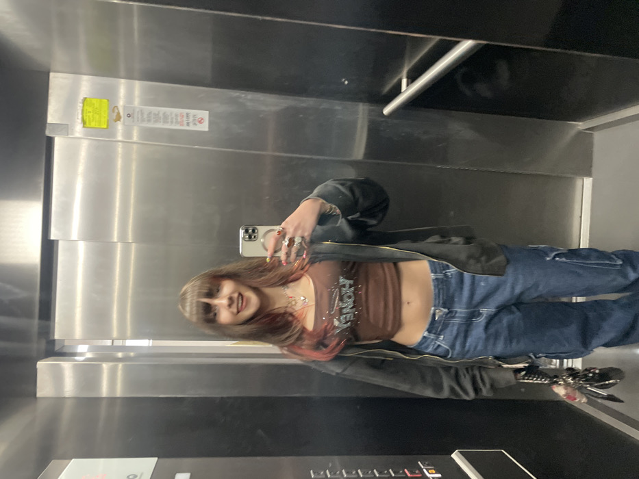 In an elevator, a young woman poses for a mirror selfie. She is wearing a brown crop top with the word "HONEY" written on it, blue jeans, and a black jacket. She has long brown hair with red highlights and bangs. Her phone is visible in her hand, and the elevator's control panel is in the foreground. The elevator is metallic and appears to be modern.