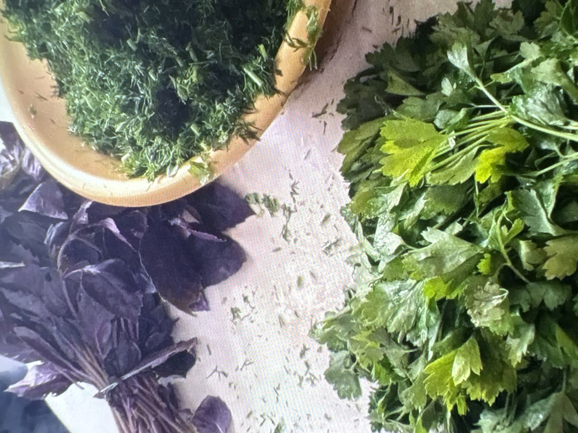In Batumi, Georgia, a close-up, high-angle shot captures an arrangement of fresh herbs on a light-colored, textured surface. To the upper left, a bunch of dark purple basil leaves, tied with a string, lies on the surface. To the right, a portion of a golden-yellow bowl contains finely chopped dill. In the foreground, a large pile of fresh, green parsley is arranged. The surface is lightly scattered with small, dried fragments of herbs. No people are visible in the scene. The lighting is soft and diffused, creating gentle shadows. The overall impression is of fresh ingredients prepared for cooking.