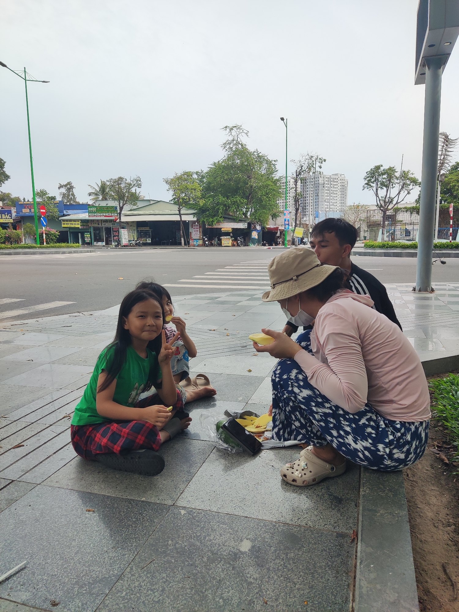 The image shows a family of four sitting on a tiled walkway next to a road with a crosswalk, in what appears to be an urban area. A young girl wearing a green shirt and plaid pants is sitting cross-legged in the foreground, holding a small piece of yellow fruit and making a peace sign with her hand. Behind her, another child sits, eating a piece of the same yellow fruit. To the right, a woman wearing a pink long-sleeved shirt, patterned blue and white pants, and a beige hat is squatting and holding a piece of yellow fruit. She is also wearing a white face mask. Behind her is a man wearing a black and white athletic jacket. Next to the woman is a dark container and pieces of yellow fruit scattered on the walkway. The road has a crosswalk and businesses are visible across the street. Streetlights and trees line the road. Tall buildings are visible in the distance.