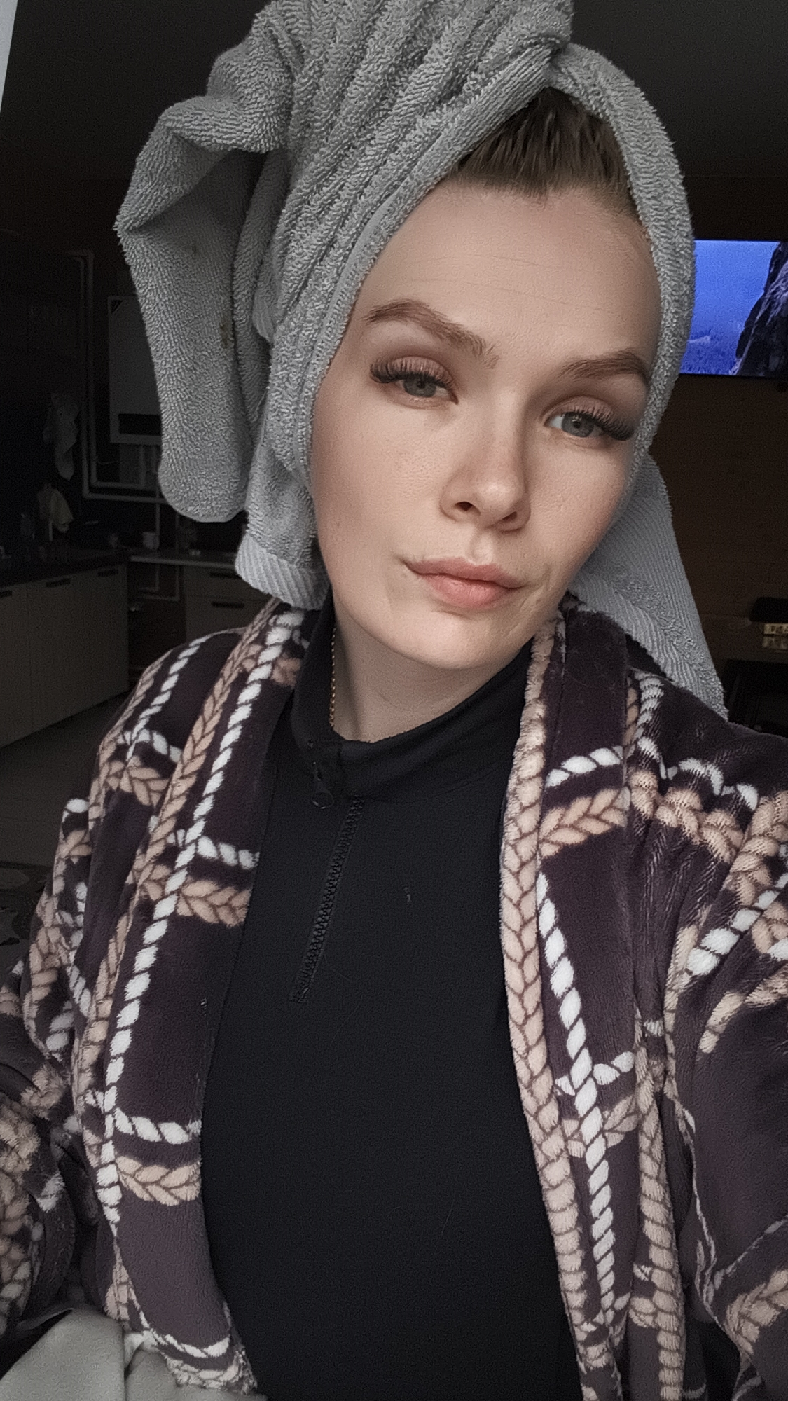 A portrait photograph features a young woman posing indoors, likely within a residential setting in Русский Вожой, Russia. She is shown from the chest up, looking directly at the camera with a calm, neutral expression. Her light hair is wrapped in a light grey terry cloth towel, suggesting she may have recently washed it. She has light-colored eyes, well-groomed eyebrows, and is wearing makeup, including visible eyelashes and subtle lip color.
She is dressed in a black high-neck top with a short front zipper, layered under a plush robe or cardigan. The robe has a distinctive patterned design featuring interlocking rope motifs in shades of white, beige, and dark brown against a darker fabric. A thin gold chain is visible around her neck, tucked beneath her black top.
The background on the left side of the image reveals glimpses of what appears to be a kitchen or utility area with light-toned surfaces. To the right, a portion of a screen, possibly a television or monitor, displays a bright blue sky over a landscape that includes what looks like a mountain or rugged terrain. The lighting is soft and consistent with an indoor environment. No text is visible in the image.