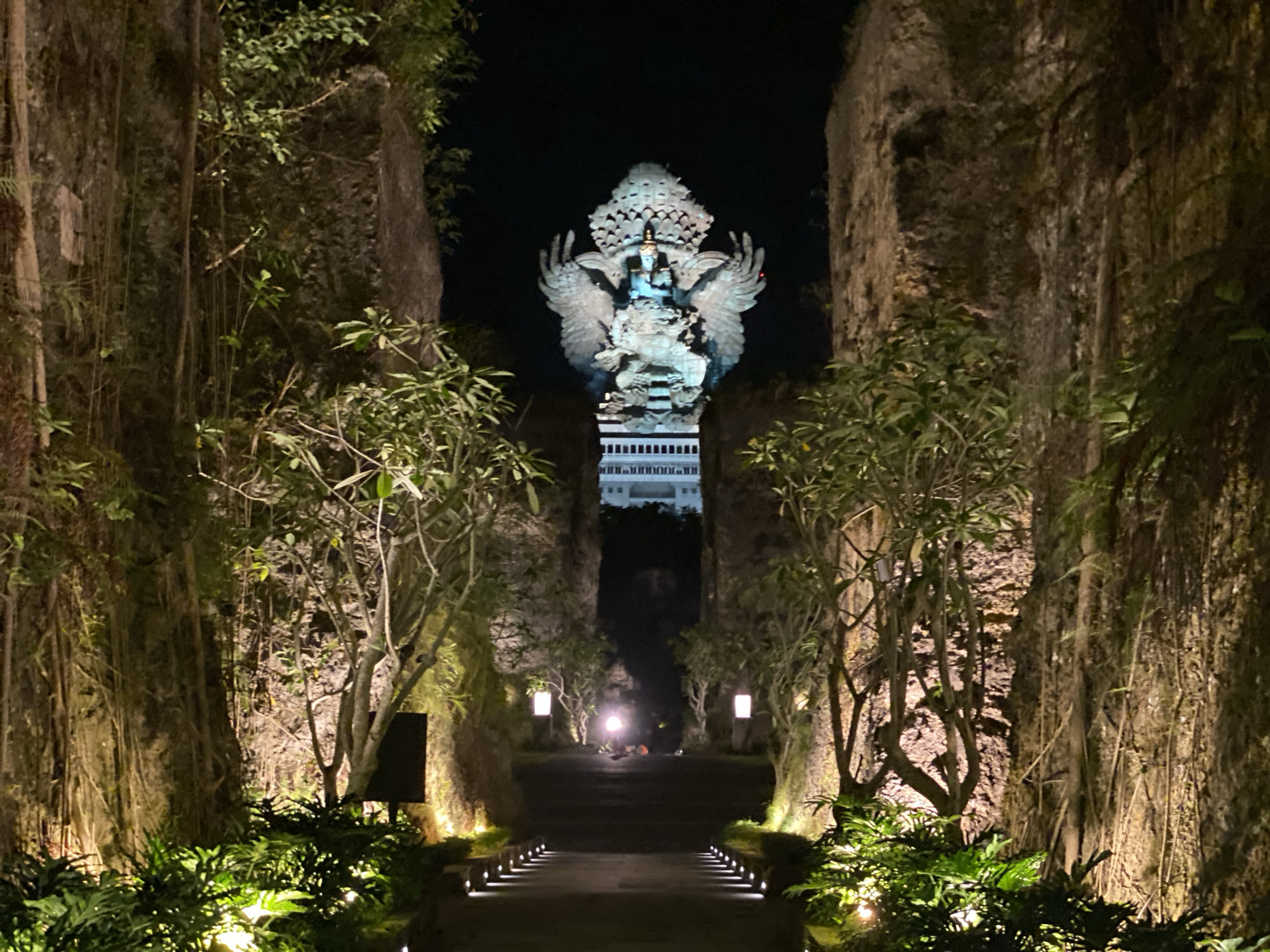 The image shows a pathway at night, leading towards a large statue of a Garuda, a mythical bird-like creature from Hindu mythology. The pathway is flanked by tall, textured rock walls covered in vegetation and illuminated by warm spotlights. The statue, centrally located and appearing to be made of bronze or a similar metal, is brightly lit, with its wings spread wide. Behind the statue, a white building with multiple floors is visible in the distance, also illuminated. The pathway itself is paved and lined with small, bright lights. Lush green plants are visible in the foreground, also illuminated by spotlights. The scene appears to be an outdoor attraction or temple at night, likely in a tropical or subtropical climate due to the vegetation.