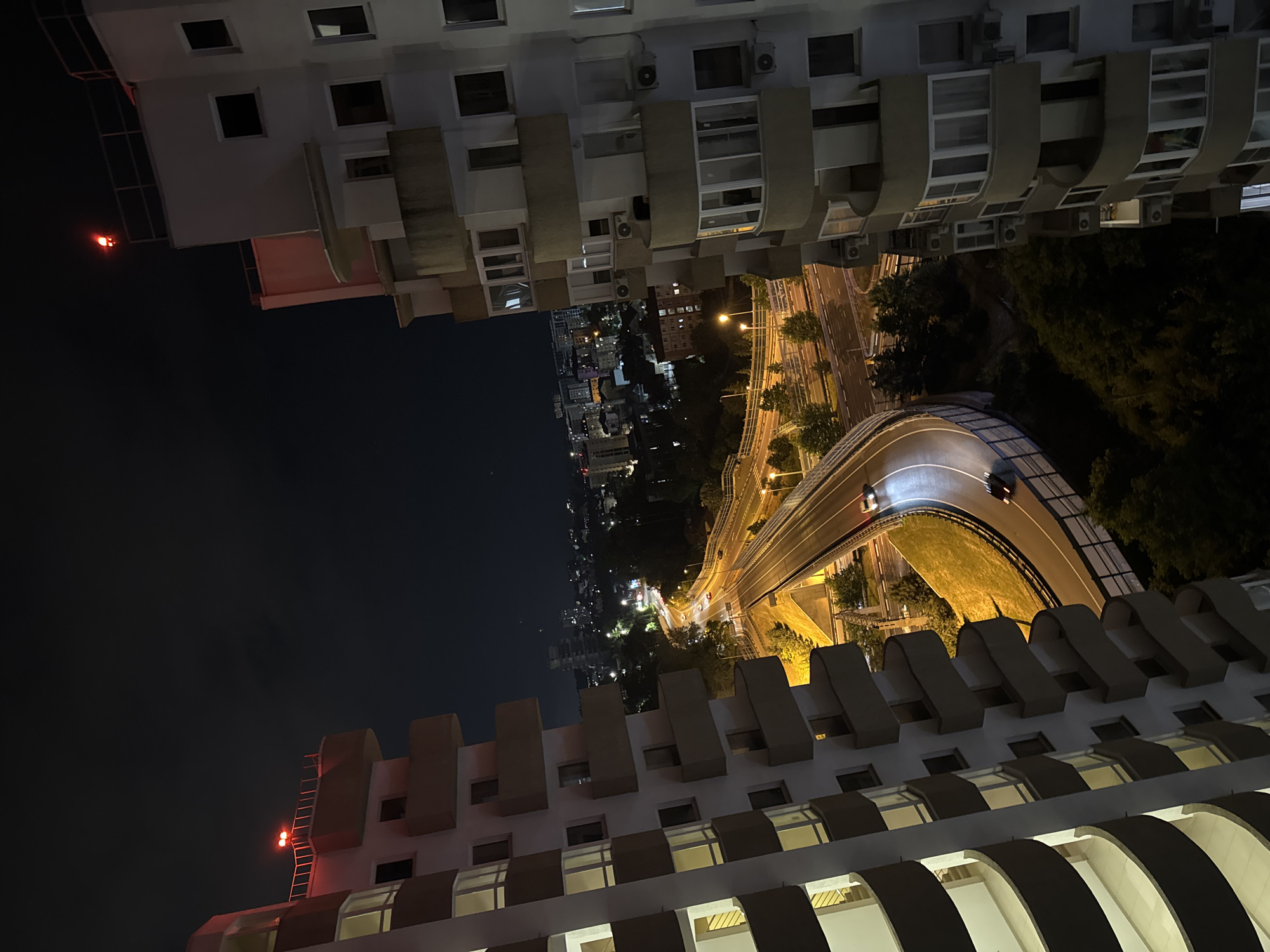 An aerial, nighttime, long shot captures a highway curving through a densely populated urban landscape in Sochi, Russia, framed by two multi-story apartment buildings. The buildings, situated on either side of the frame, feature curved balconies and illuminated windows. Red aviation lights are visible on the rooftops of both buildings, indicating their height. The highway is well-lit by streetlights, with two vehicles visible on the road: a white car and a darker car. The surrounding area is filled with numerous buildings of varying heights, illuminated by interior lights and streetlights, characteristic of a city at night. Trees are scattered throughout the landscape, particularly denser in the foreground, adding to the urban greenery. The sky is dark, with no visible stars or moon, suggesting a cloudy or overcast night.