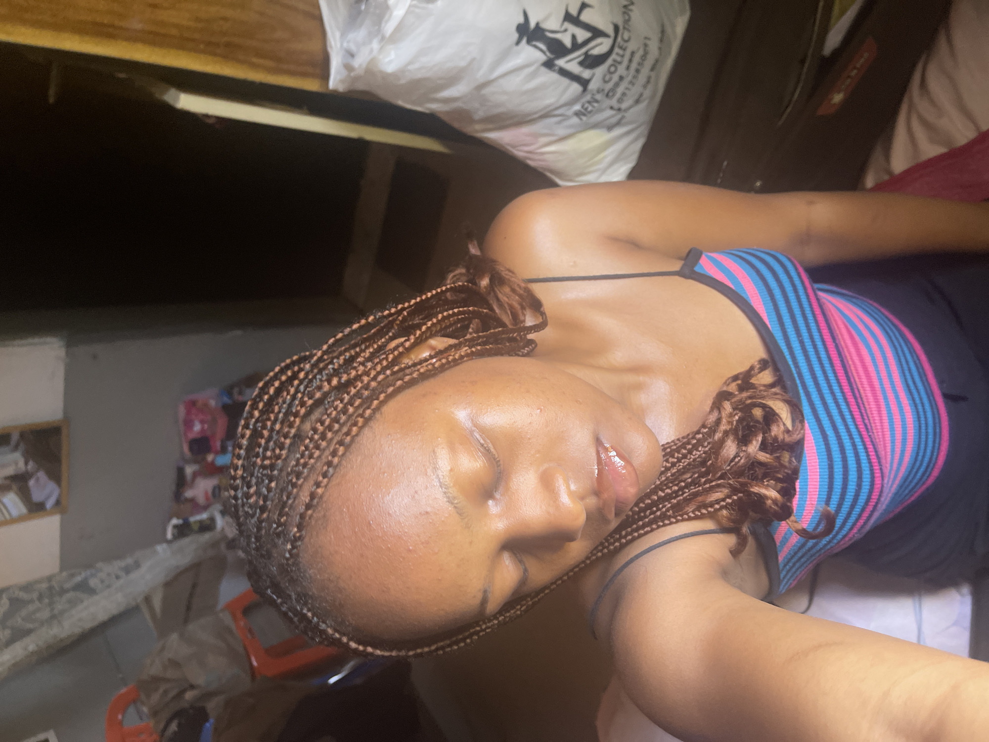 In an indoor setting, possibly a bedroom in Rafin Par, Nigeria, a young woman with braided hair is captured in a close-up, overhead shot. She is wearing a striped tank top in pink, blue, and black, and her eyes are closed. Her skin appears slightly shiny, suggesting it may be humid or she may have applied a product. The background is cluttered with various items, including what appears to be a large white bag with writing on it, a wooden surface, and possibly furniture. The lighting is dim, casting shadows and highlighting the contours of her face. The overall scene conveys a sense of personal reflection or relaxation.
