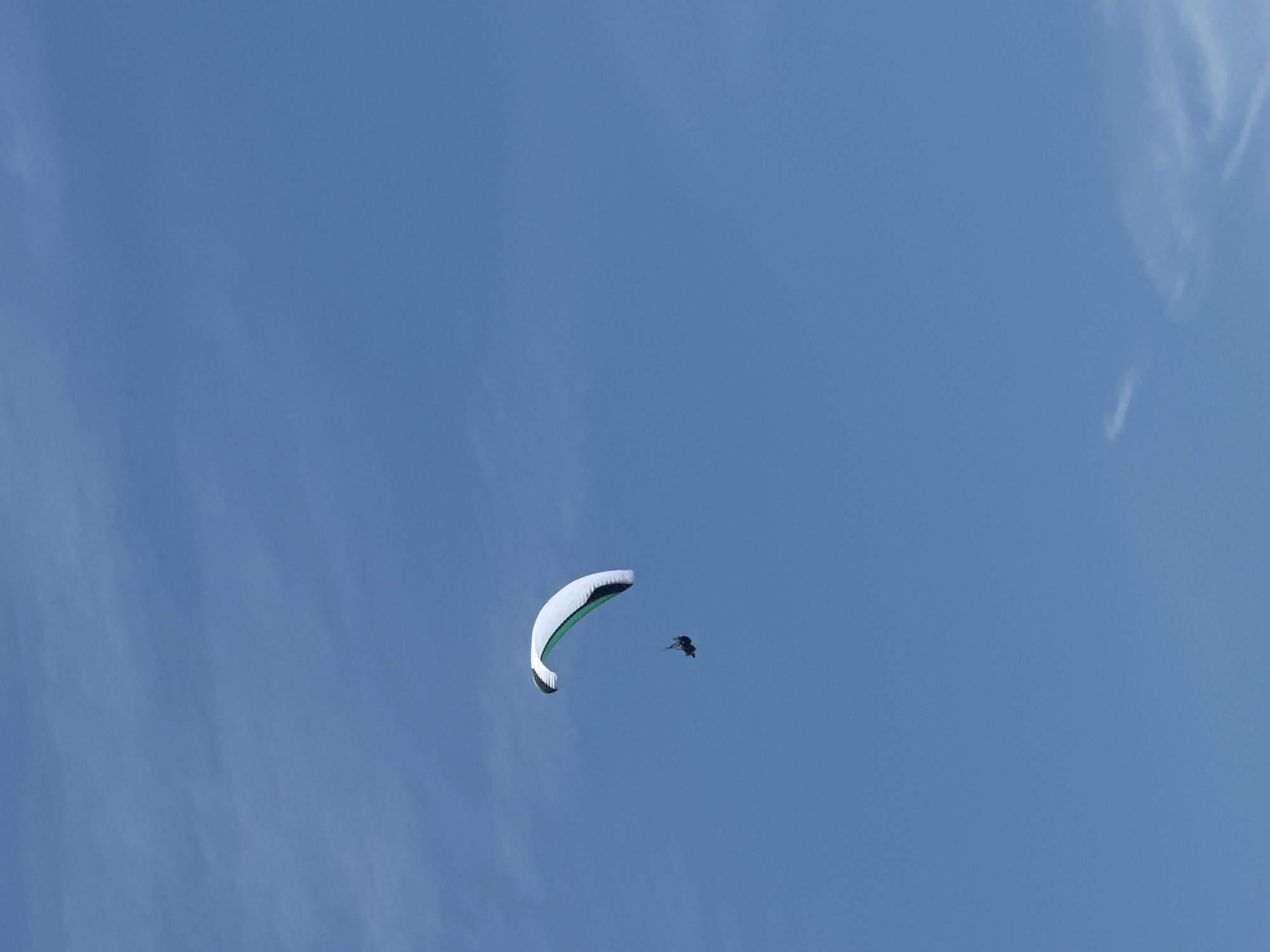 A paraglider is seen in the sky over Boreti, Montenegro. The paraglider has a white wing with a green stripe. A single person is suspended beneath the wing in a harness. The sky is a clear blue with a few wispy clouds. The paraglider appears to be at a considerable altitude. The image captures a moment of leisure activity against a backdrop of natural beauty.