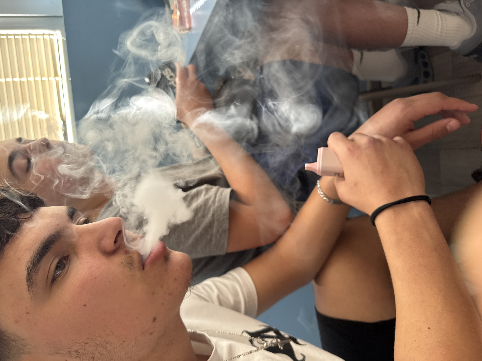 In Yambol, Bulgaria, two individuals are depicted indoors, engaged in vaping. The foreground is dominated by a young male exhaling a plume of vapor. He is wearing a white t-shirt with a silver necklace and black shorts. To his right, another individual, partially obscured by the vapor, is wearing a grey t-shirt and blue shorts. Both individuals are seated, with blinds visible in the background. The scene is captured in a candid, close-up perspective, emphasizing the act of vaping and the resulting vapor.