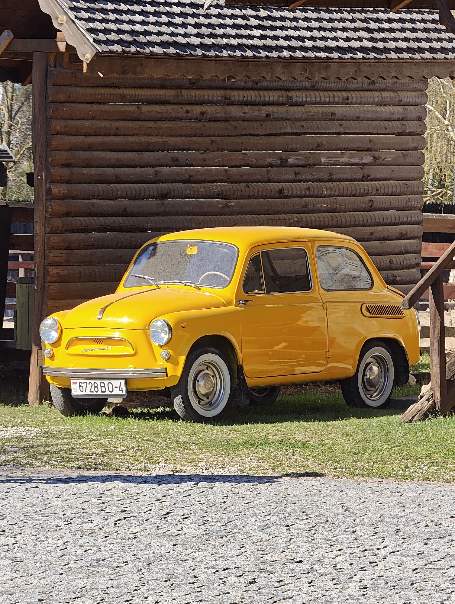A yellow ZAZ-965 car is parked on a patch of grass beside a small log cabin-style structure. The car has white-walled tires and a license plate that reads "6728 BO-4" with the country code "BY". The location is Капцёўскі сельскі Савет, Belarus. The foreground shows a paved area. The log structure has a dark shingle roof.