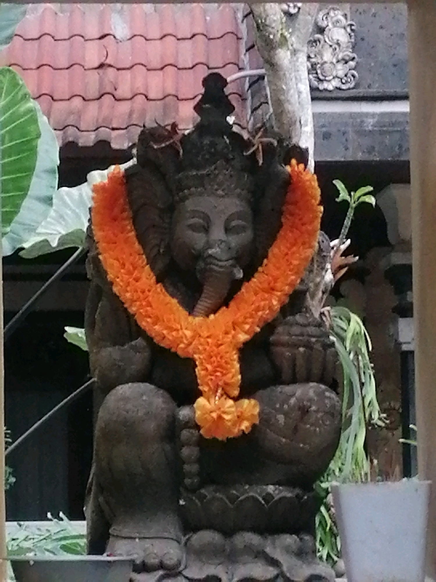 In Ubud District, Indonesia, a stone statue of Ganesha, adorned with a vibrant orange flower garland, is centrally positioned. The statue sits on a tiered base with lotus petal carvings. Behind the statue, a rustic tiled roof is visible, and to the right, a grey stone wall with decorative carvings and a small potted plant with green leaves. The scene is framed by wooden elements on the sides, suggesting an outdoor setting, possibly a temple courtyard or garden.