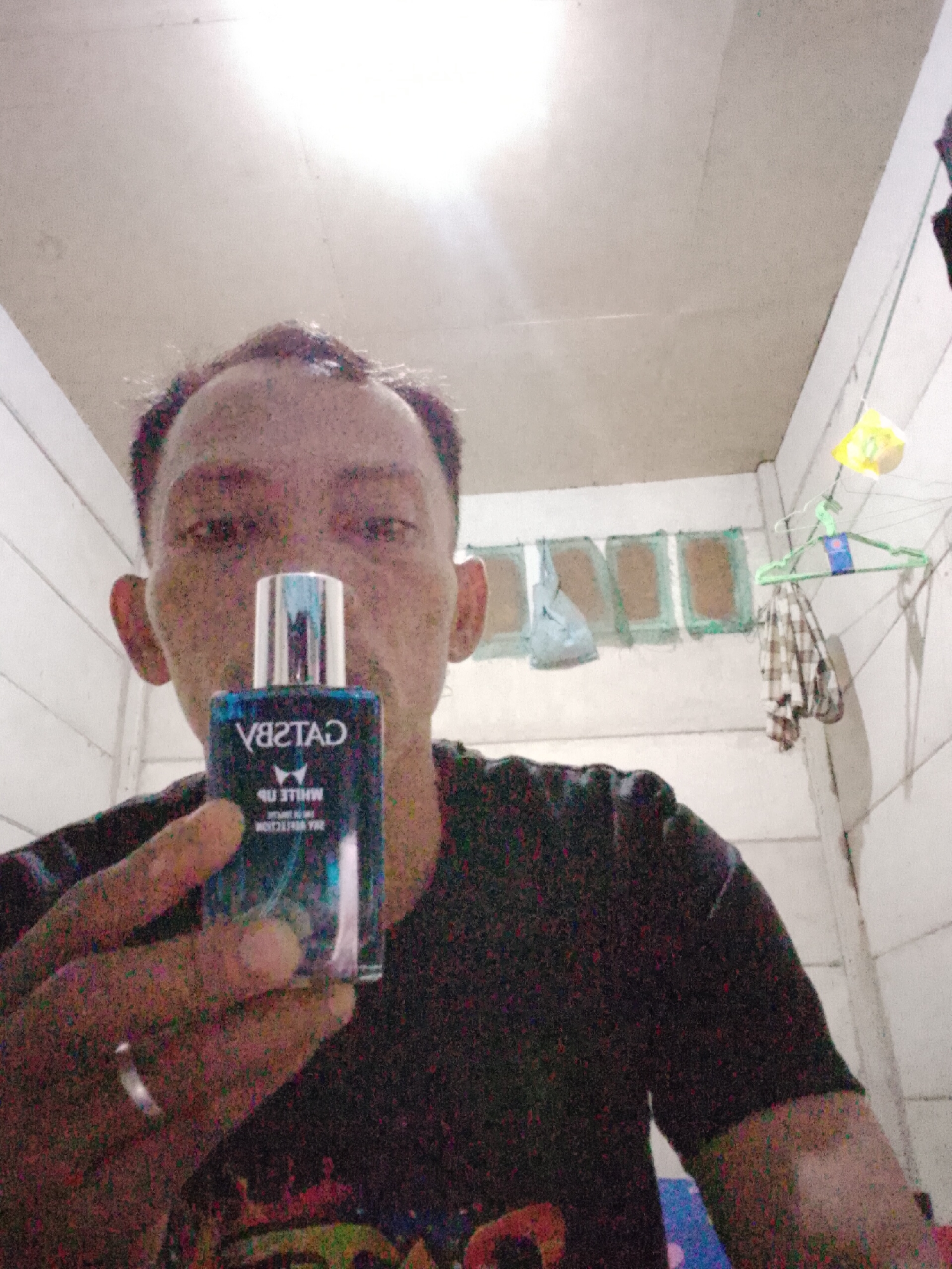 A man is holding up a blue bottle of Gatsby White Up deodorant spray, with the label facing the camera and appearing reversed. He is wearing a black t-shirt and a ring on his left hand. The background consists of a white wooden wall and ceiling, with a clothesline strung across the wall. There are some items hanging on the clothesline, including a green hanger with a plaid shirt. The lighting suggests an indoor environment. The image is grainy and slightly out of focus, typical of a low-quality photo.